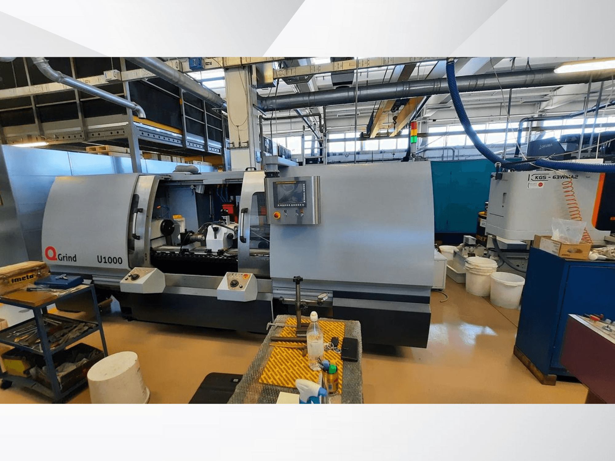 Q-Grind U1000 mašīna darbnīcā, priekšējais skats ar vadības paneli un atklāto griešanas zonu, ko ieskauj iekārtas.