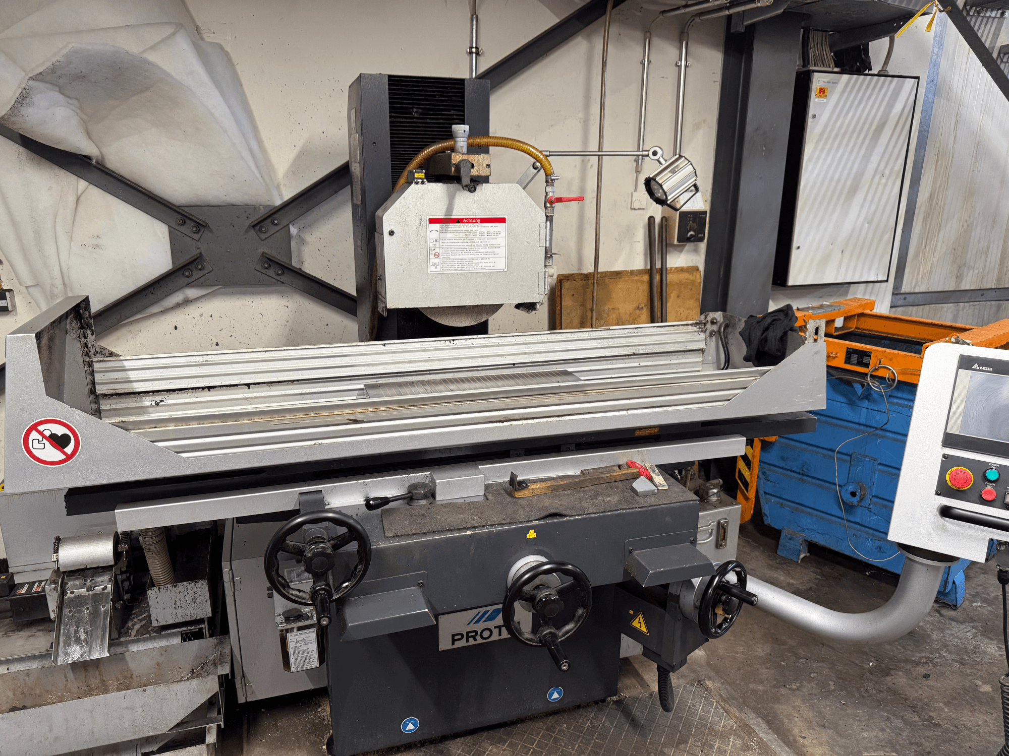 PROTEC mašīna, priekšējais skats, kurā redzama virsmas slīpmašīna ar manuālo vadību, drošības brīdinājumiem un dzesēšanas sistēmu.