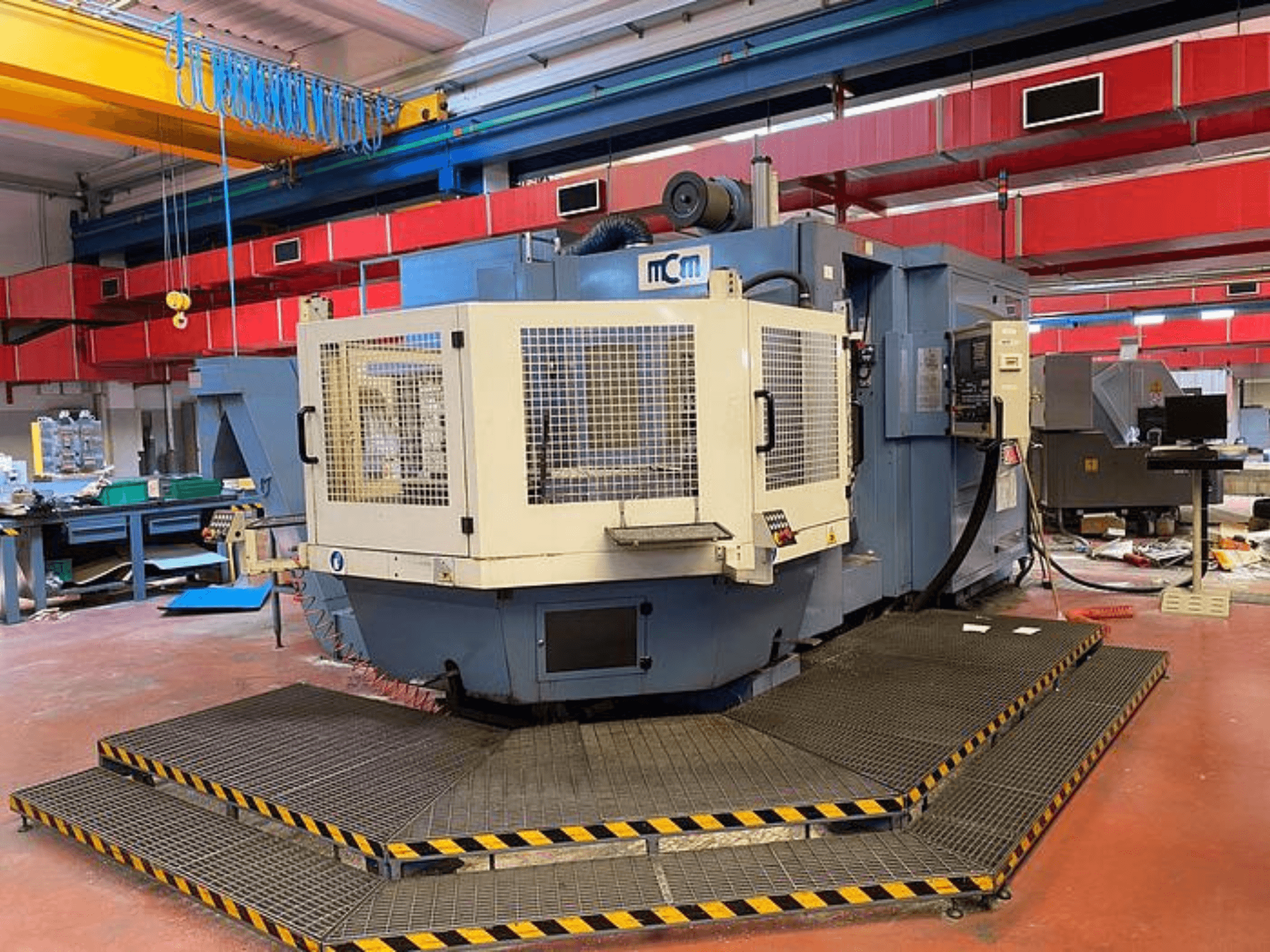 MCM CNC darbgalds rūpnīcā, skats no priekšpuses ar aizsargapvalku, melnas un dzeltenas krāsas drošības apmalēm, ieskauj rūpnieciskās iekārtas.