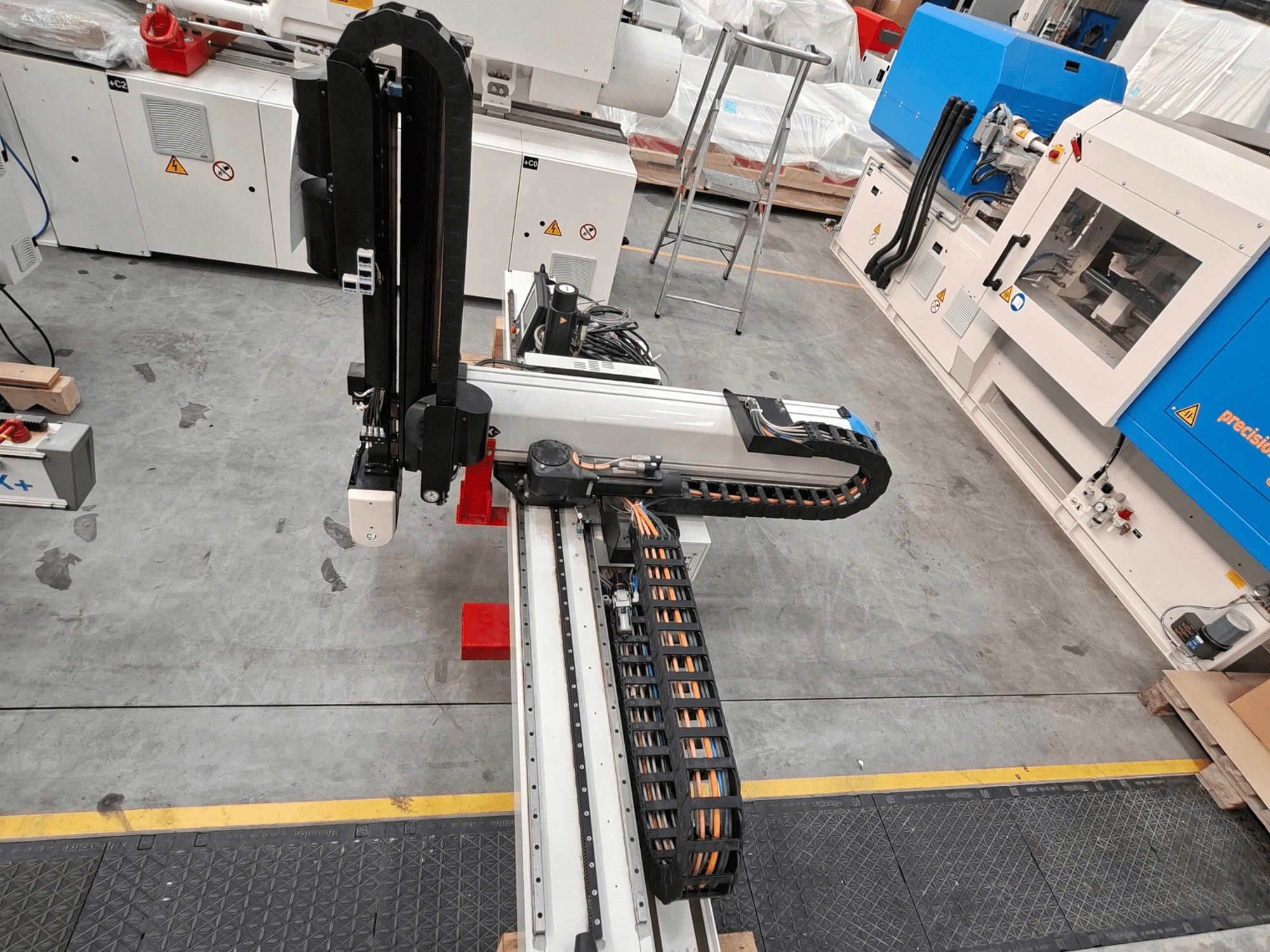 Plastmasas pārstrādes iekārta „Krauss Maffei LRX150-MC5” skatā no augšas, kurā redzama robota roka un kabeļu trase ražotnes grīdā.