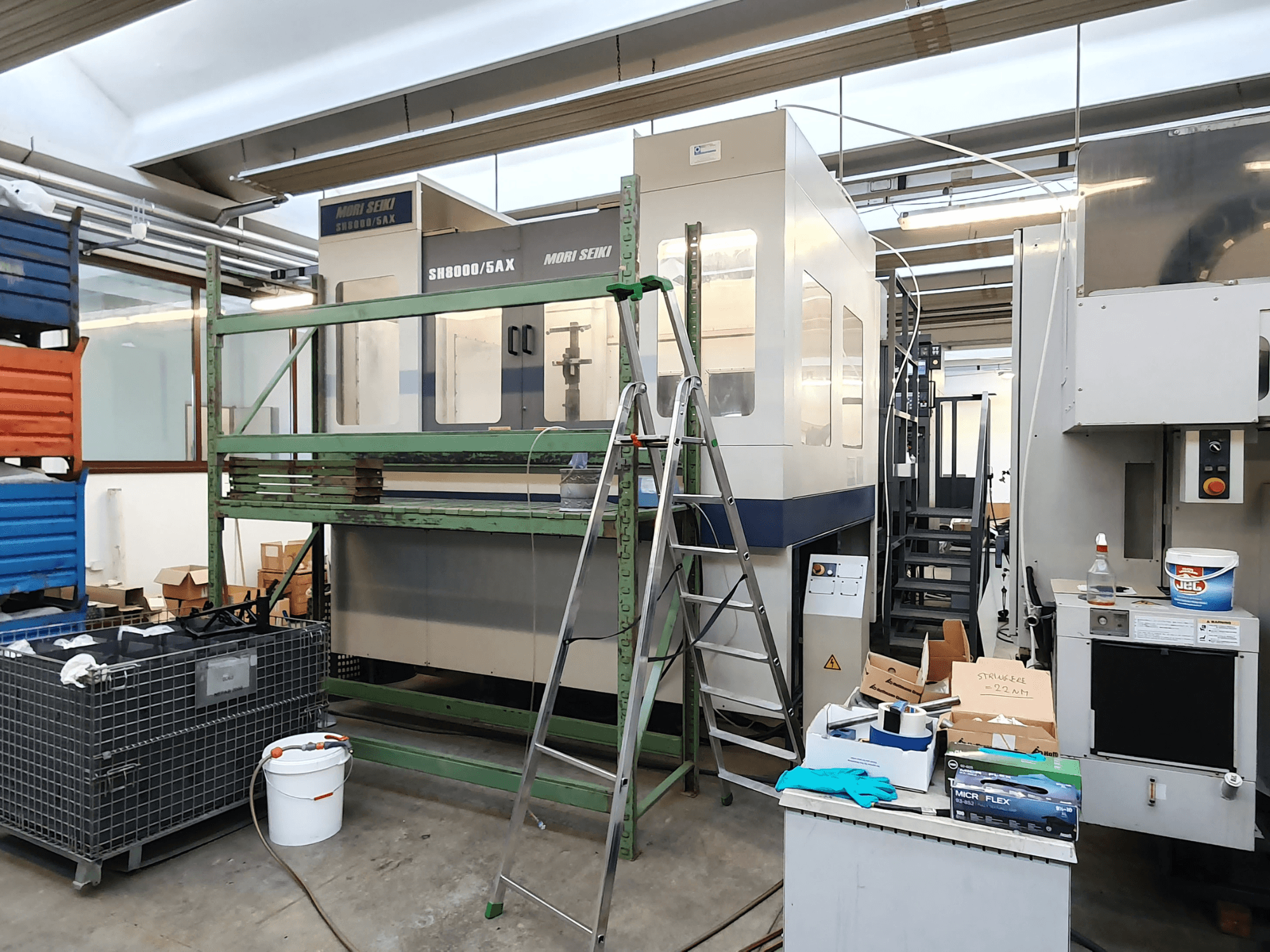 Mori Seiki SH-8000/5AX CNC darbnīcā, skats no priekšpuses, ar darbnīcas instrumentiem un glabāšanas kastēm blakus.