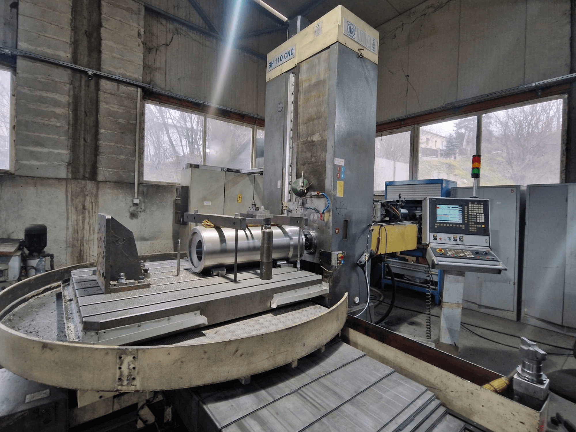 LOLA BH 110 CNC urbjmašīna darbnīcā, skats no priekšpuses, ar metāla sagatavi un vadības paneli.