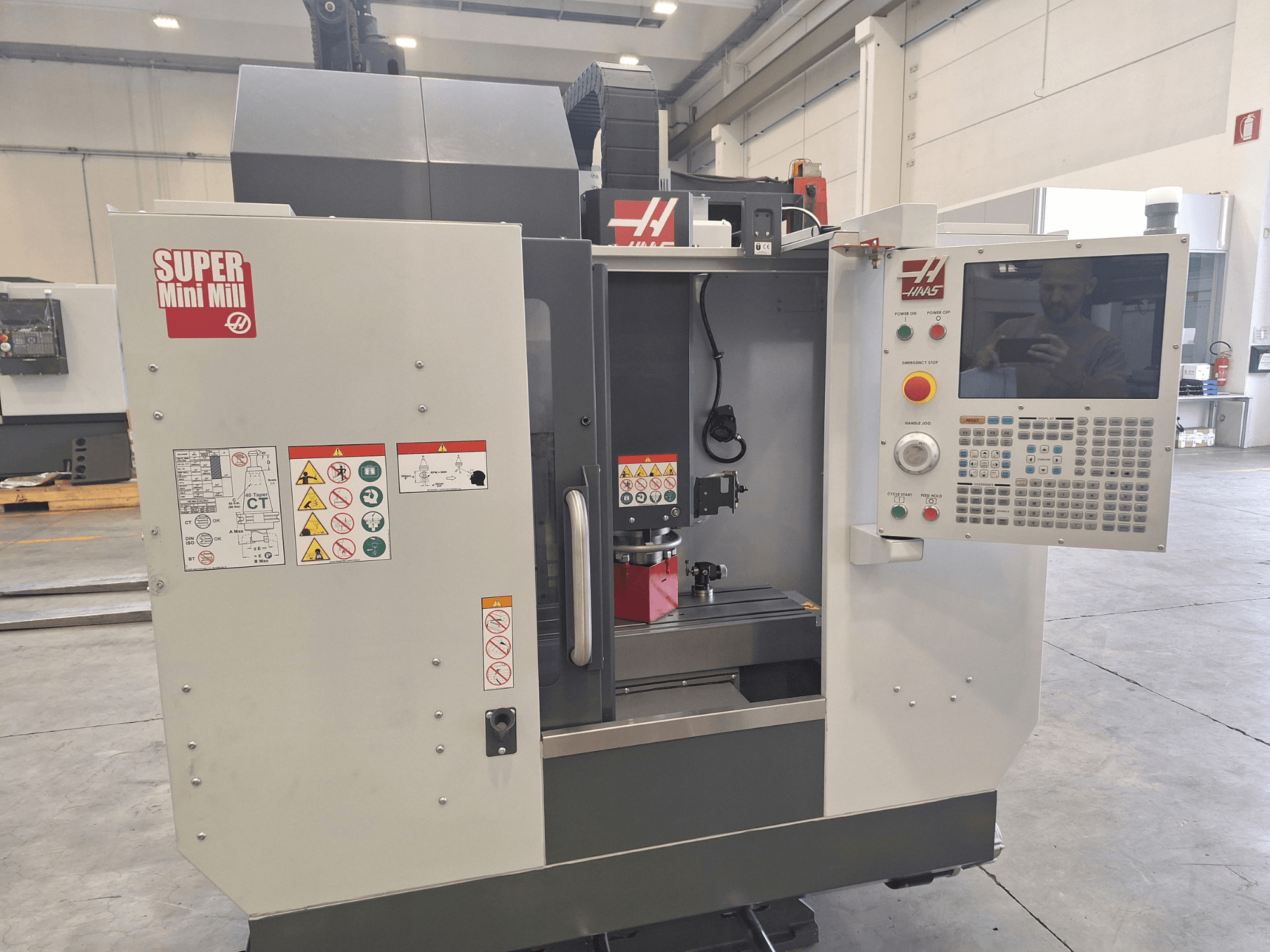 HAAS Super Mini Mill skats no priekšpuses, kurā redzams vadības panelis, drošības uzlīmes un apstrādes zona ar uzstādītu detaļu.