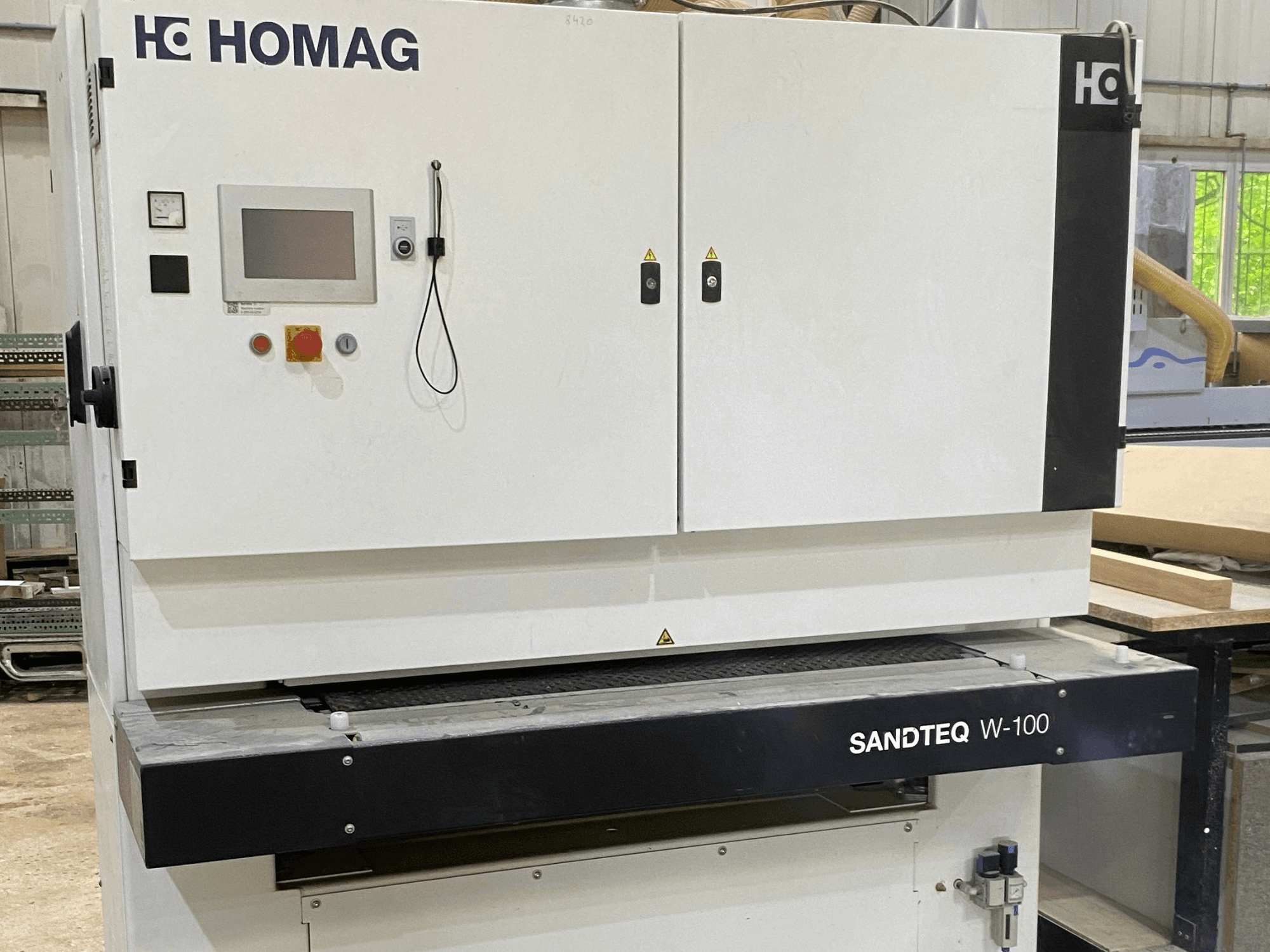 HOMAG SANDTEQ W-100 lentes slīpmašīna darbnīcā, kurā redzams vadības panelis, putekļu nosūces atvere un slīpēšanas lentes zona. Skats no priekšpuses.