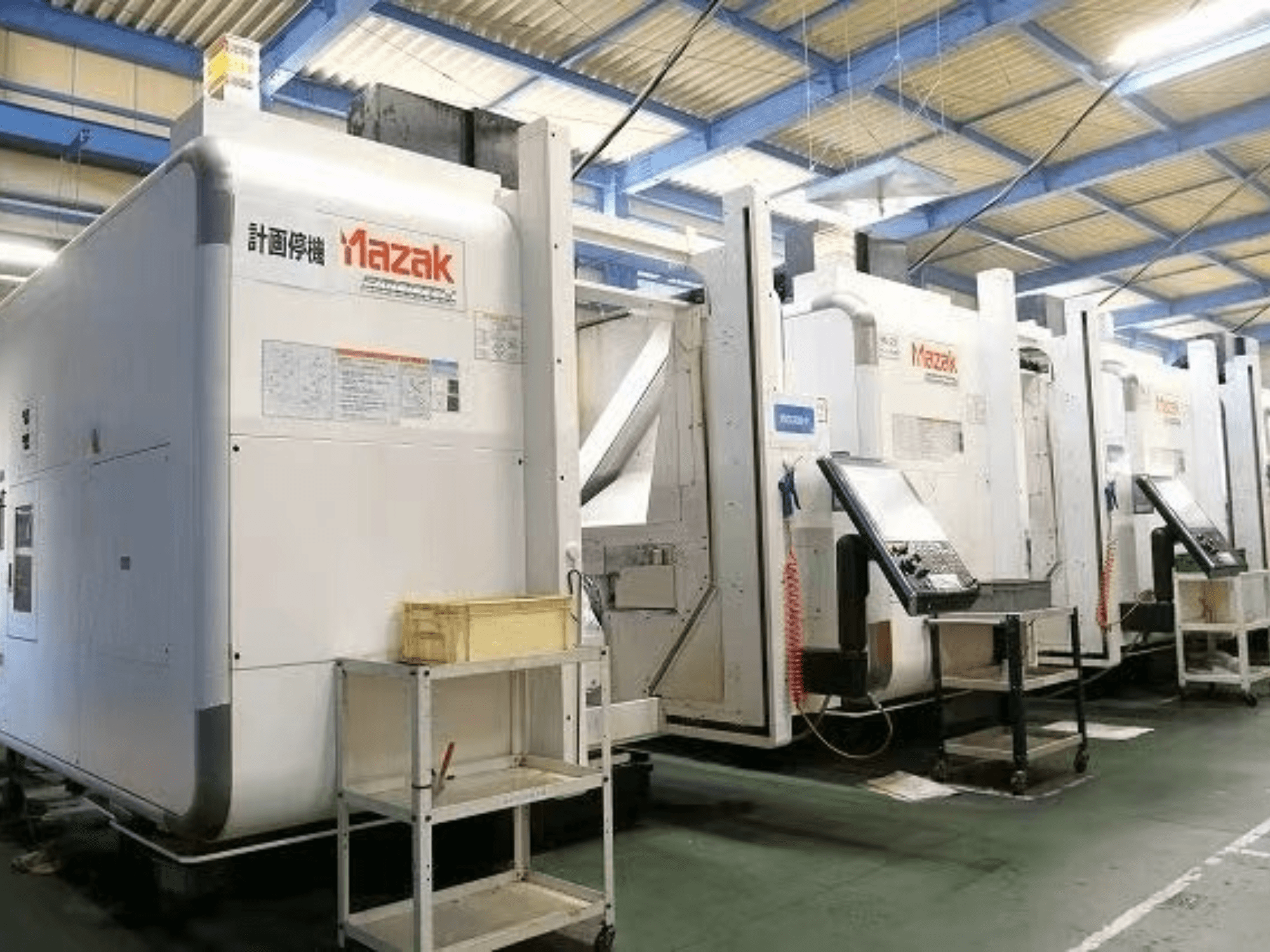 Mazak CNC darbgaldi rūpnīcā, skats no sāniem, kur redzami vadības paneļi un etiķetes uz baltiem korpusiem.