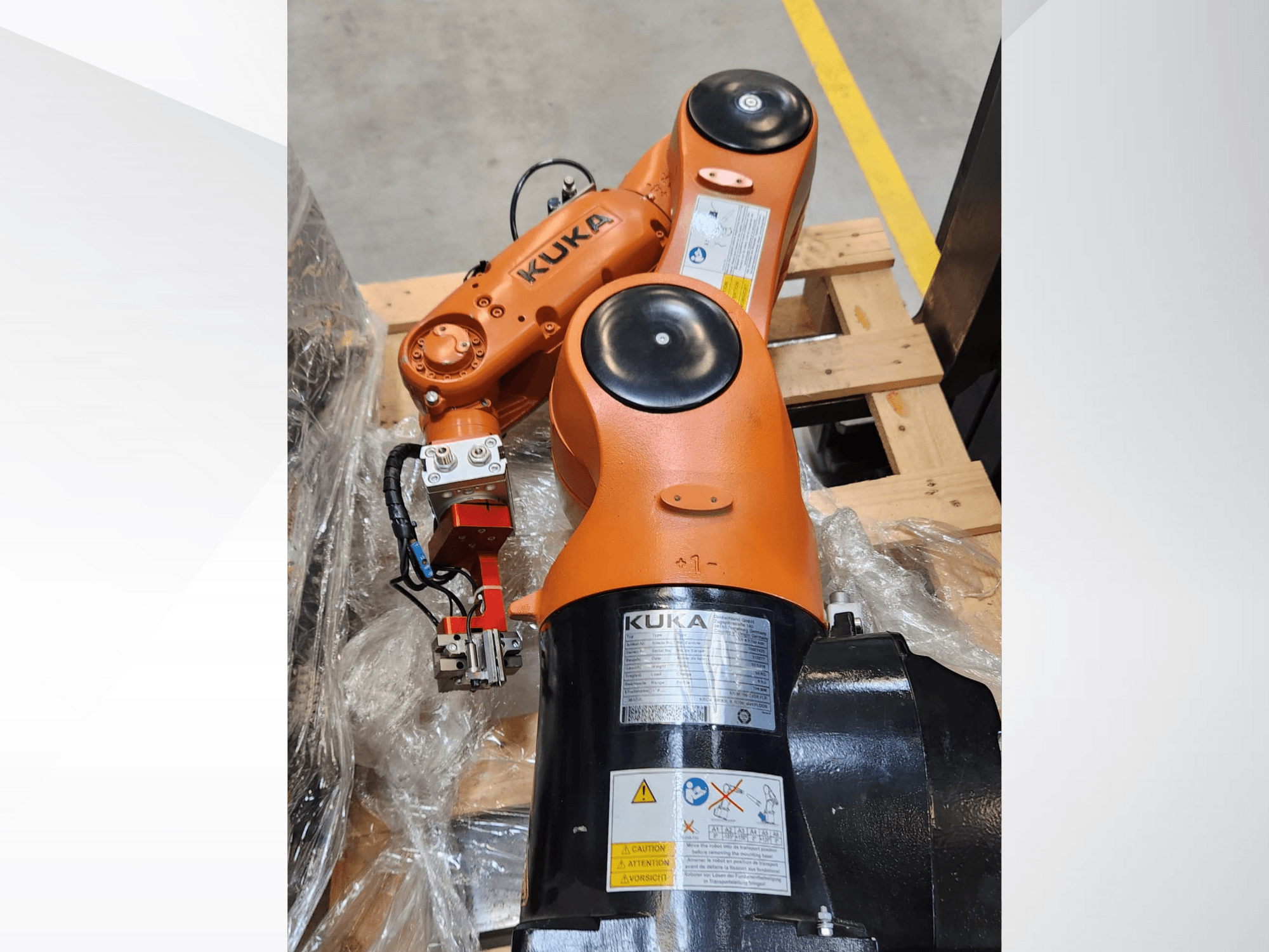 KUKA robotizētā roka ar oranžu apdari, skats no sāniem, ar satvērēju un vadības vadiem uz koka paletes.