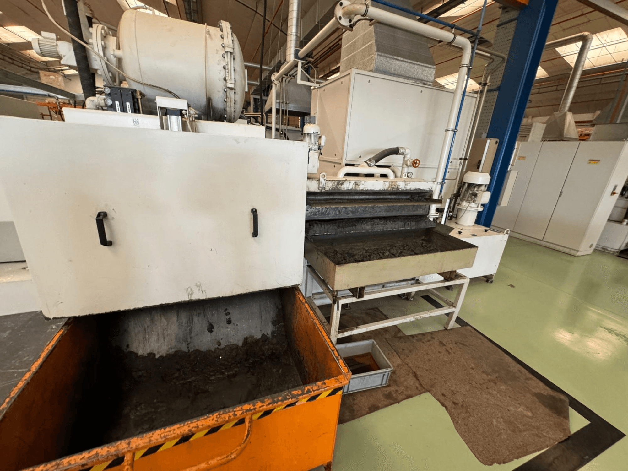 Rūpnieciskās apstrādes iekārta ar oranžu atkritumu tvertni, skats no priekšpuses; tajā ir motors, konveijeri un apkopes zona.