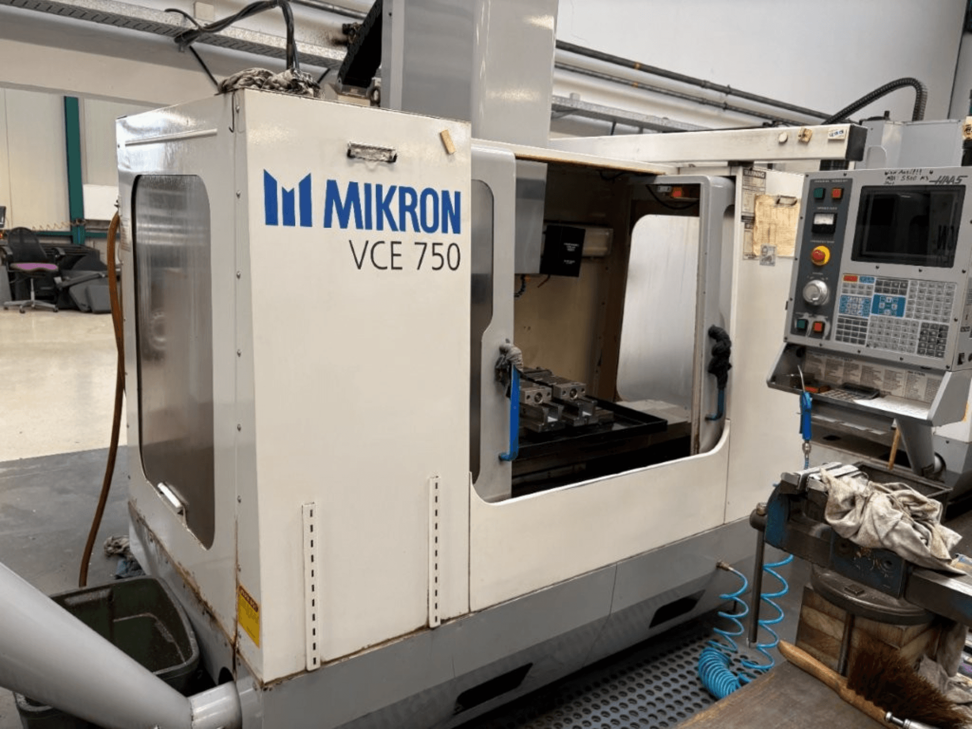Mikron VCE 750 CNC frēzmašīnas priekšējais skats ar apstrādātu darba galdu un vadības paneli ar dažādām pogām un ekrāniem.