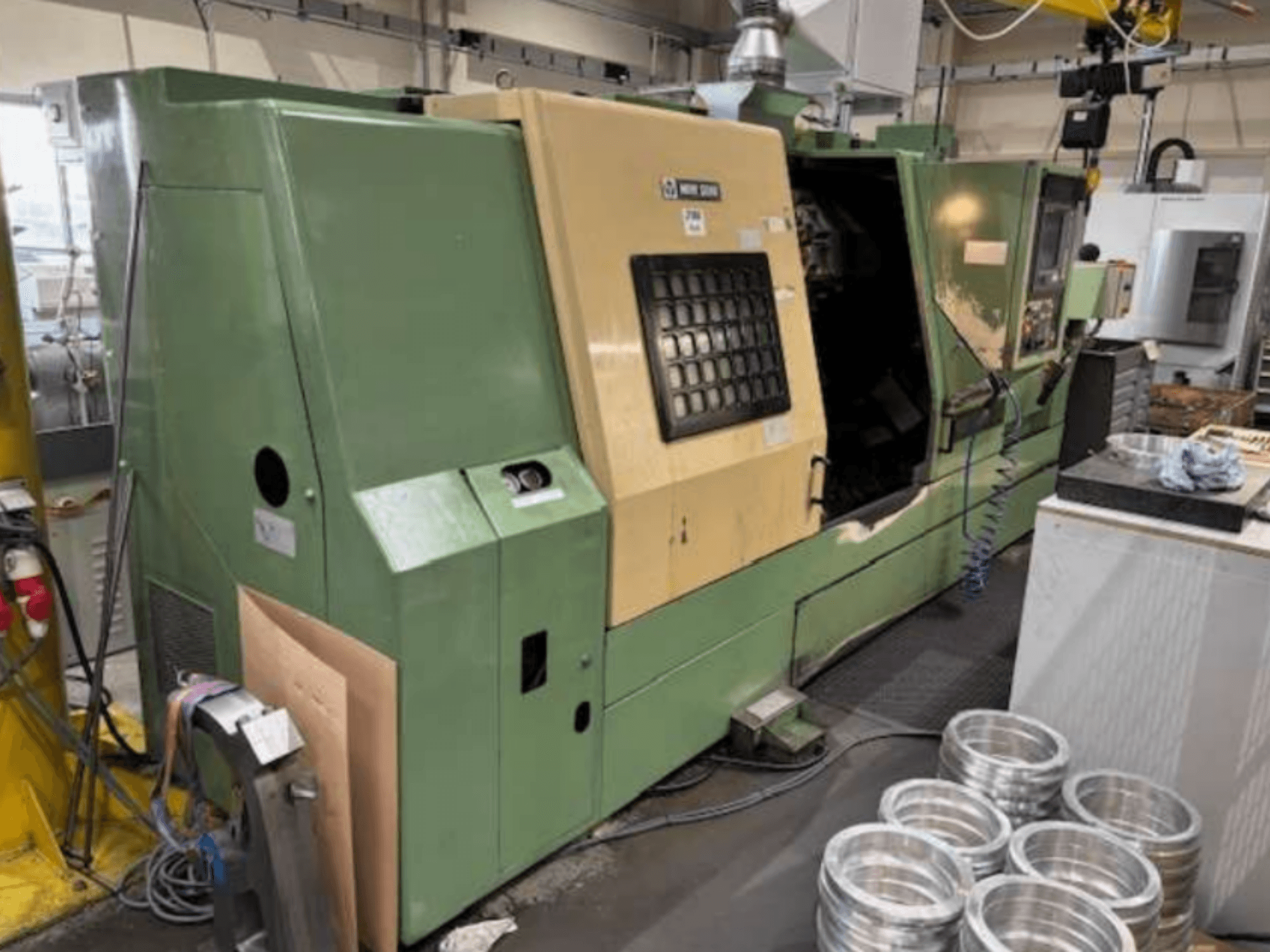 Zaļās Mazak CNC virpas, skatoties no priekšpuses, ar vadības paneli un apstrādātām alumīnija detaļām uz darba galda.