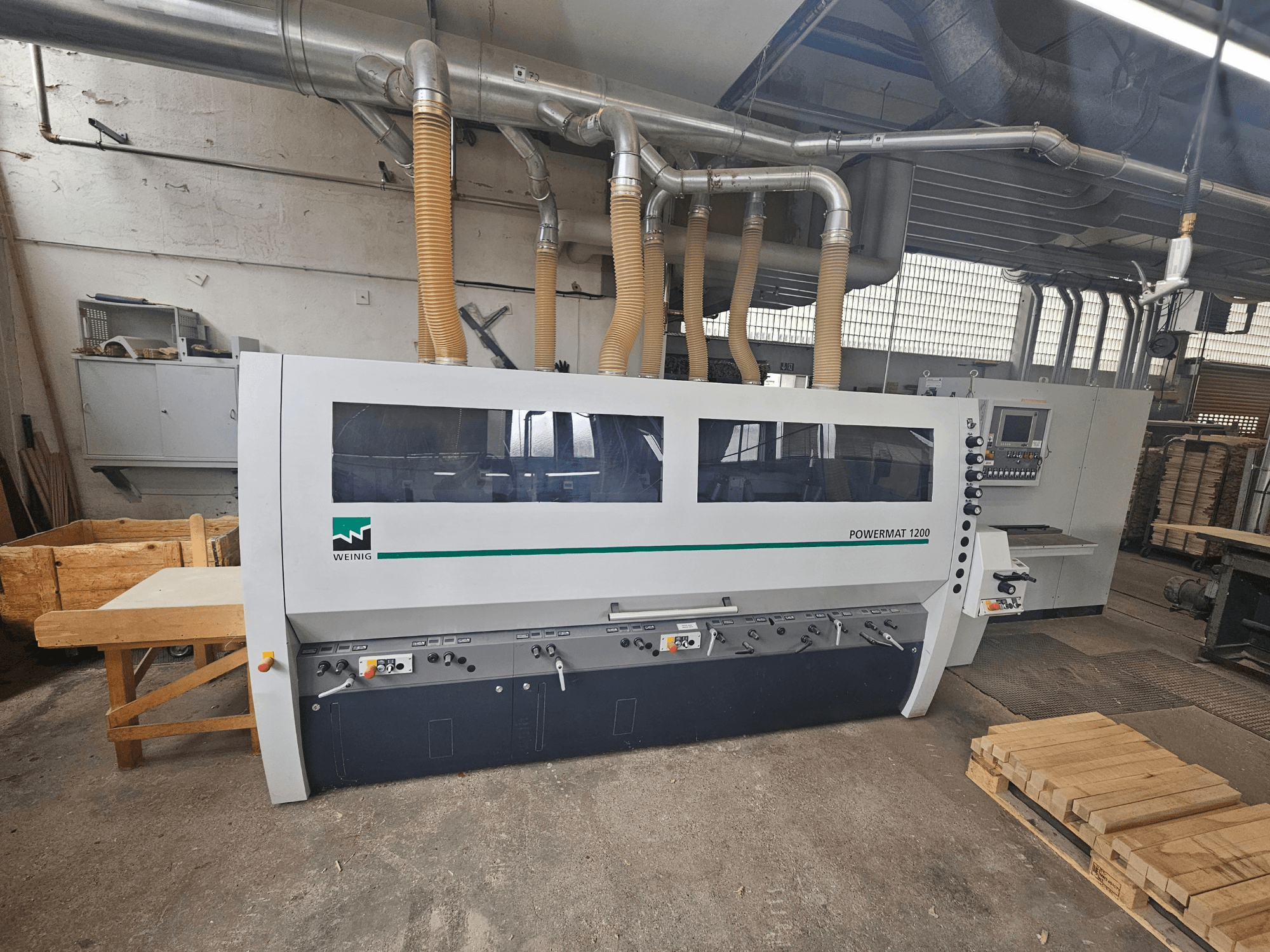 Weinig Powermat 1200 kokapstrādes darbagalds, skats no priekšpuses, ar vadības paneli un koka kaudzēm darbnīcā.