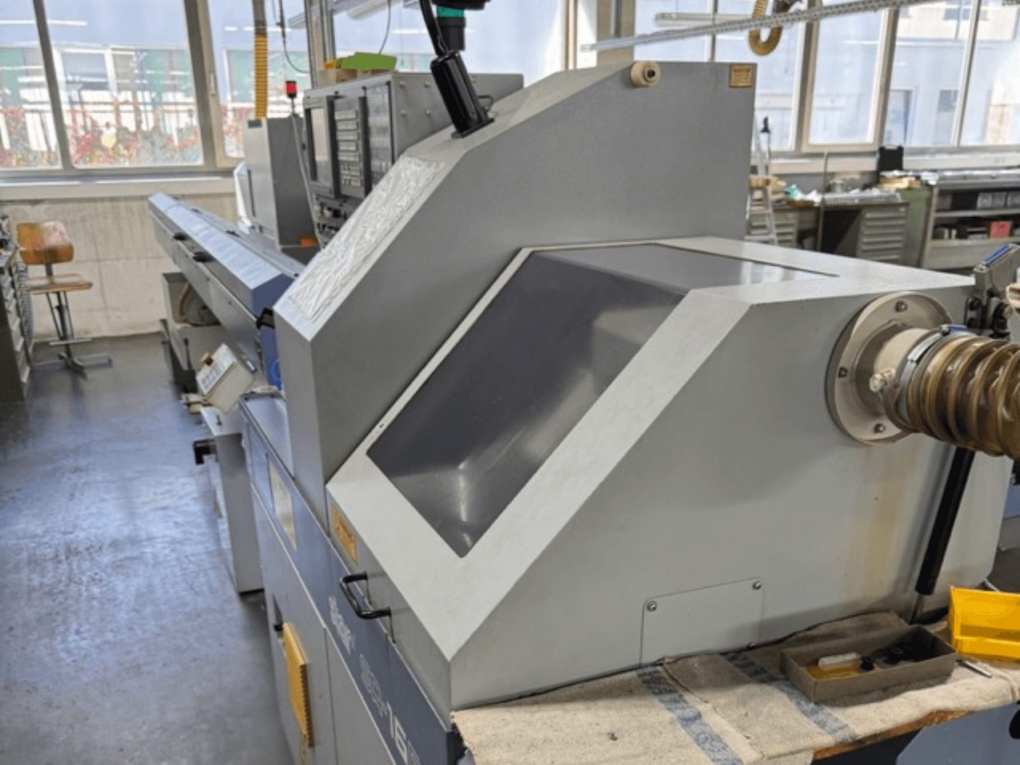 Pelēka industriāla mašīna, skatoties no sāniem, ir aprīkota ar lielu leņķveida paneli, vadības pogām un šļūtenes stiprinājumu.