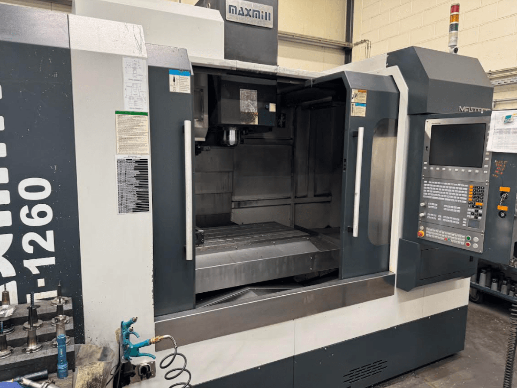 Maxmill CNC frēzmašīna, modelis M-1260, priekšējais skats, kurā redzama tukša darba vieta ar vadības paneli un lietošanas instrukcijām.