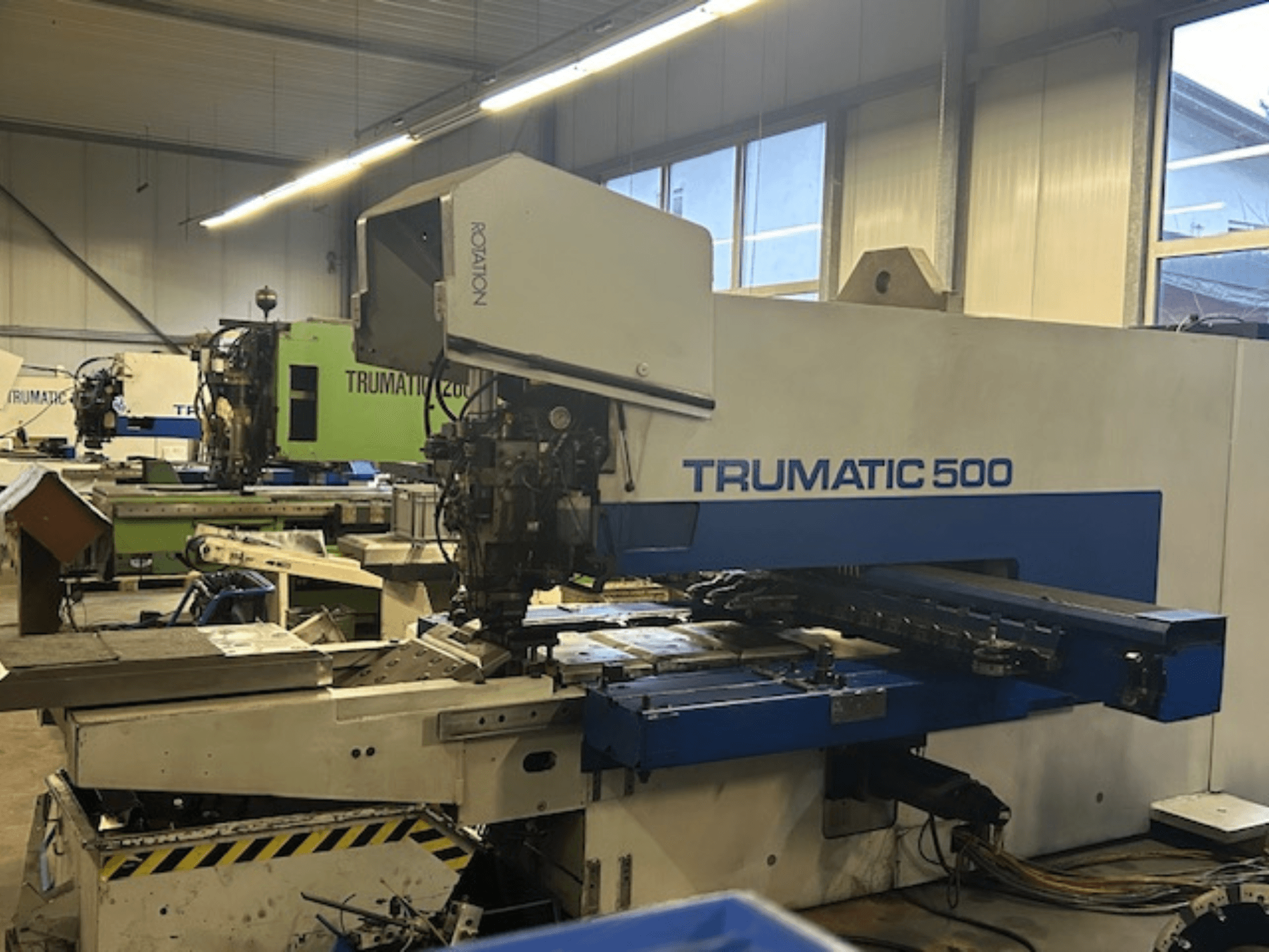 TRUMPF TRUMATIC 500 CNC caurumošanas iekārta, skats no priekšpuses, ar vadības paneli un mehāniskajām daļām. Rūpnieciskās darbnīcas vide.