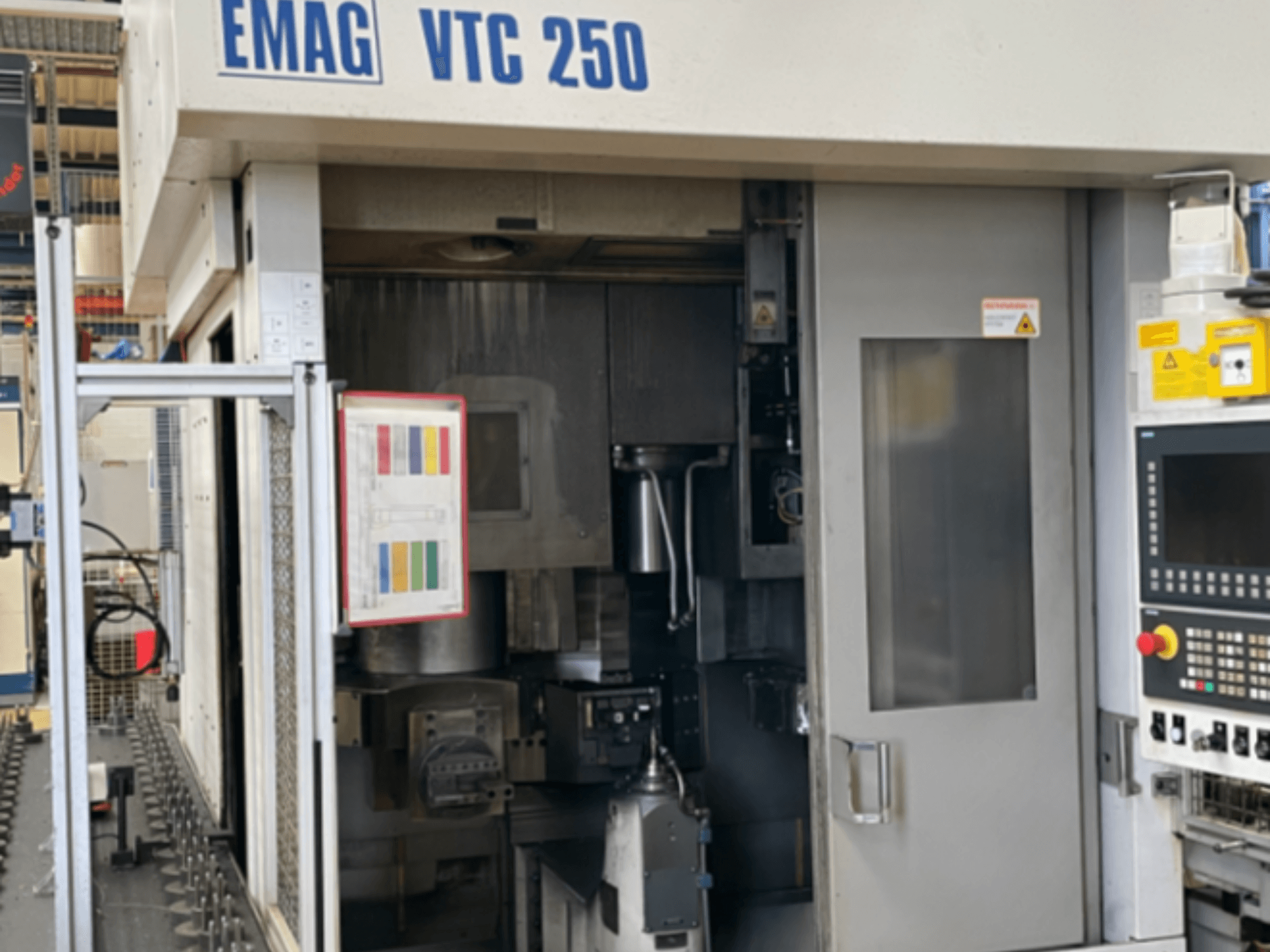 EMAG VTC 250 vertikālās virpošanas mašīnas skats no priekšpuses, kurā redzams vadības panelis, lietotāja interfeiss un atvērtās piekļuves durvis.