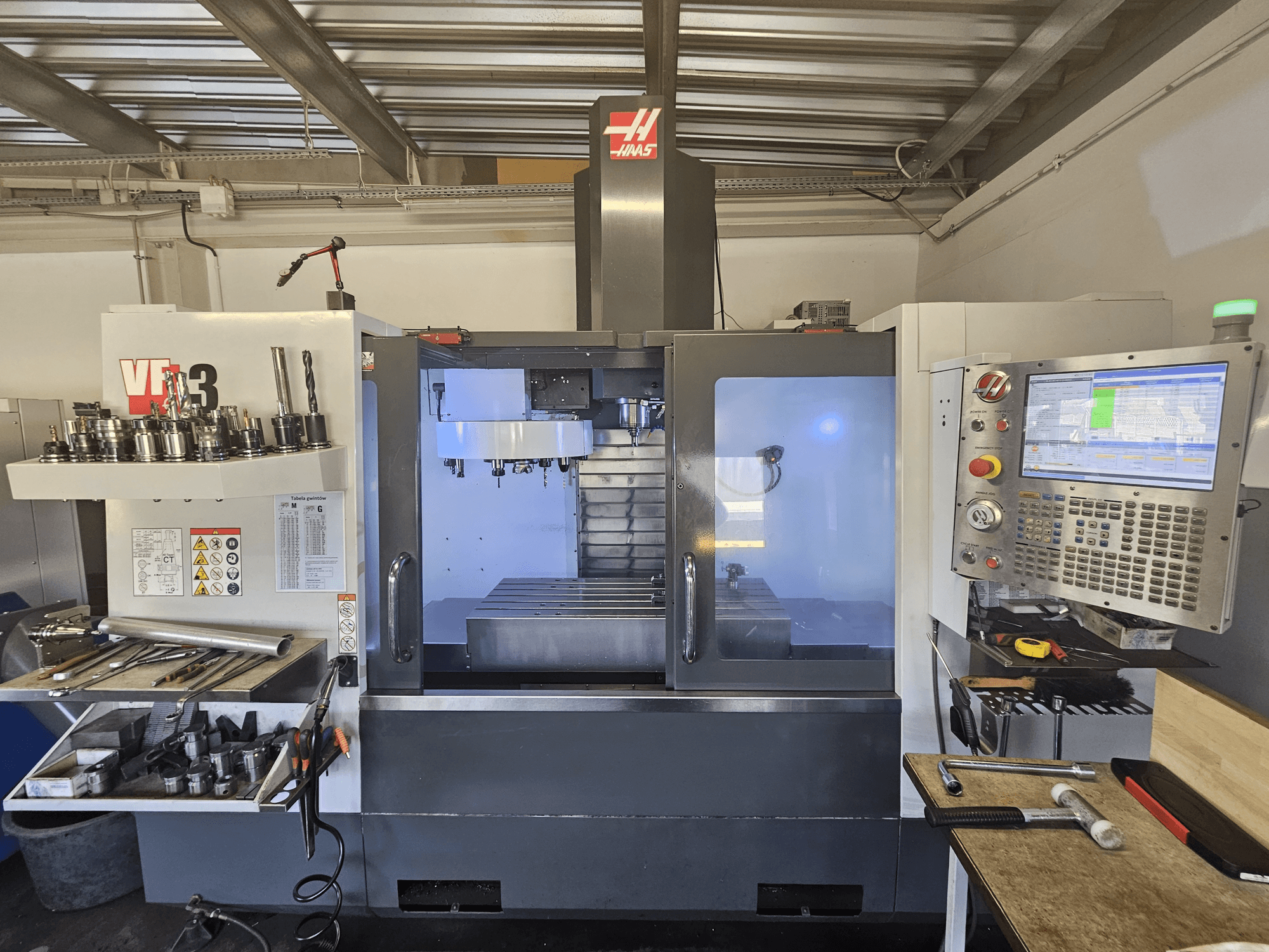 Haas VF-3 CNC darbnīcā, priekšējais skats, kurā redzami instrumenti, vadības ierīces un frēzēšanas zona ar apgaismotu iekšpusi.