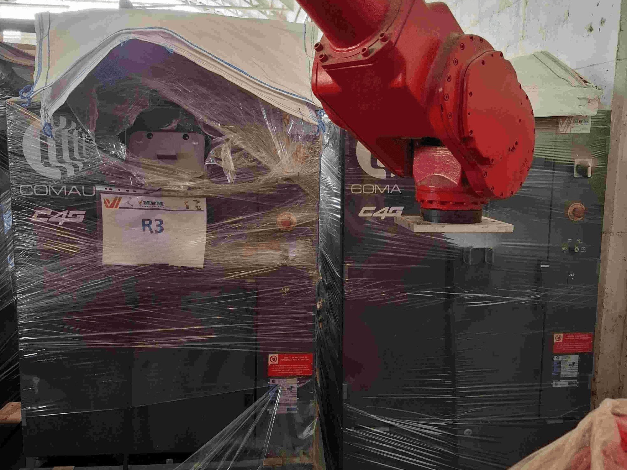 Comau C4G robotizētā roka no priekšpuses, ietīta plastmasas apvalkā, ar marķējumu "R3", uz iekārtas redzamas brīdinājuma uzlīmes.