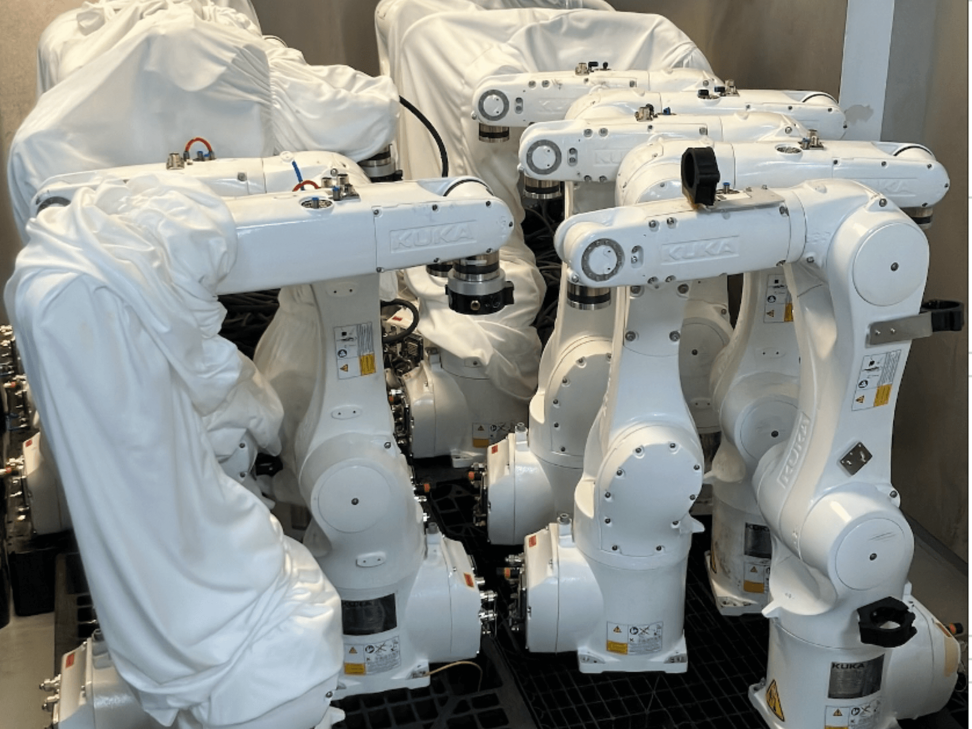 KUKA KR 10 R900-2 robota rokas, kas pārklātas ar baltu audumu, skatīts no augšējā leņķa, redzamas vairākas vienības noliktavā.