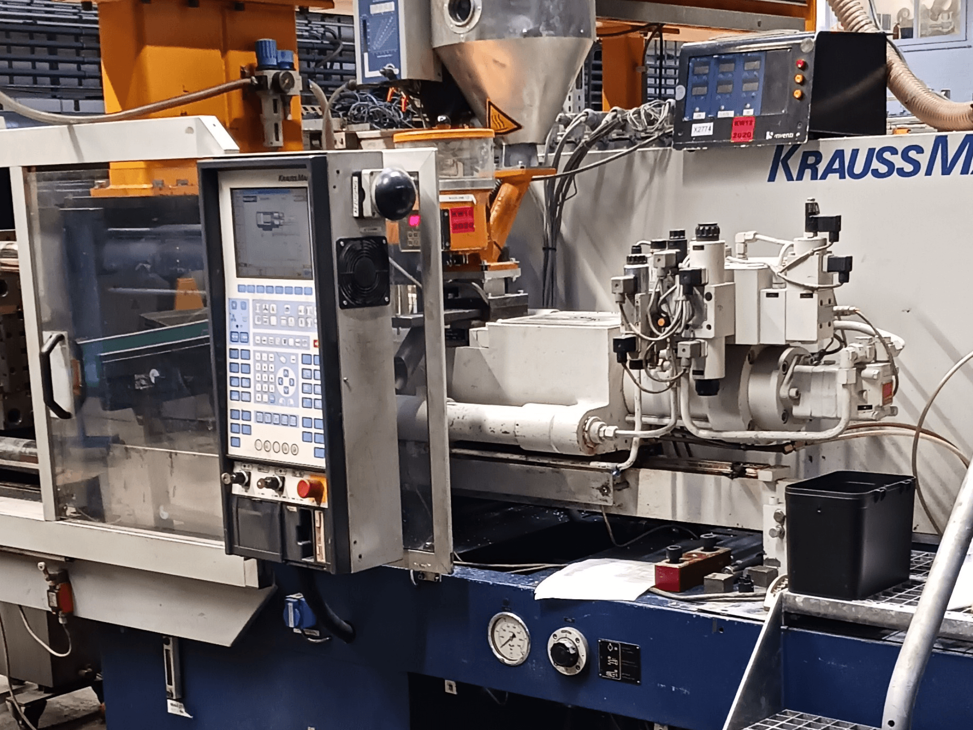 KraussMaffei iesmidzināšanas liešanas iekārta ar vadības paneli, hidrauliskajiem agregātiem un instrumentiem priekšplānā, skats no priekšpuses.