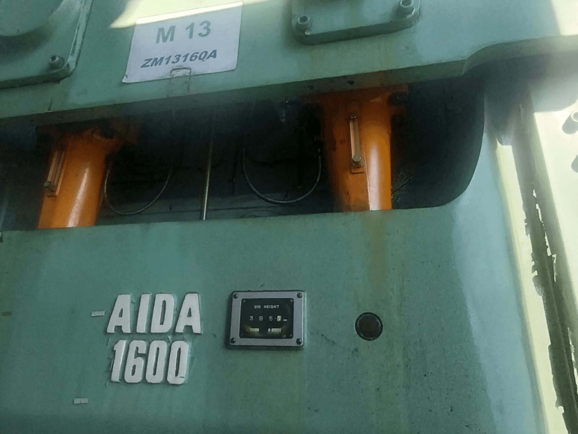 AIDA NC2-1600 mehāniskā štancēšanas prese, skats no priekšpuses; redzami oranži hidrauliskie komponenti un preses augstuma mērītājs.
