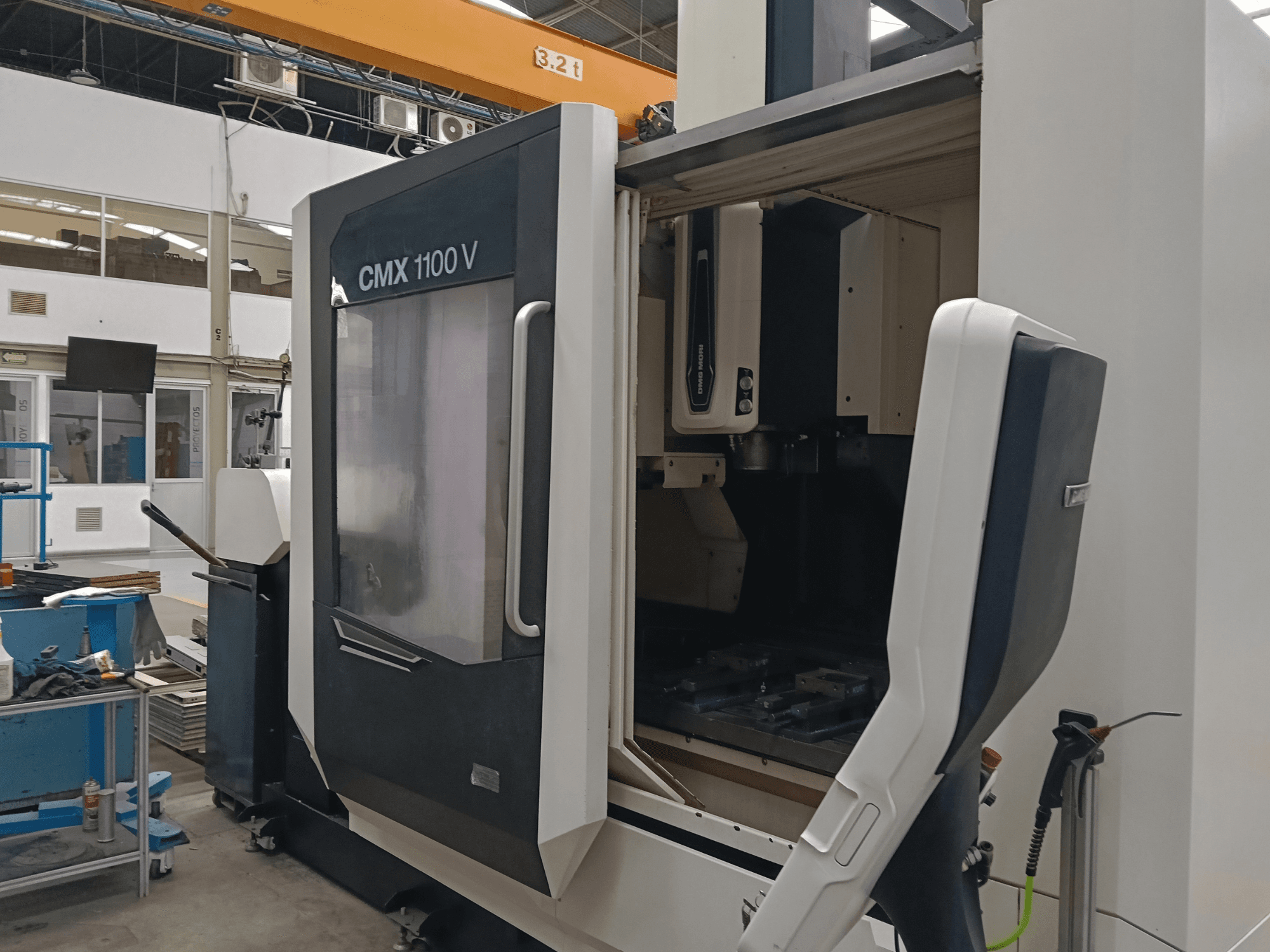 Vertikālais apstrādes centrs DMG MORI CMX 1100 V, skats no priekšpuses, kurā redzamas atvērtas durvis un iekārtas iekšējā instrumentu konfigurācija rūpniecības telpā.