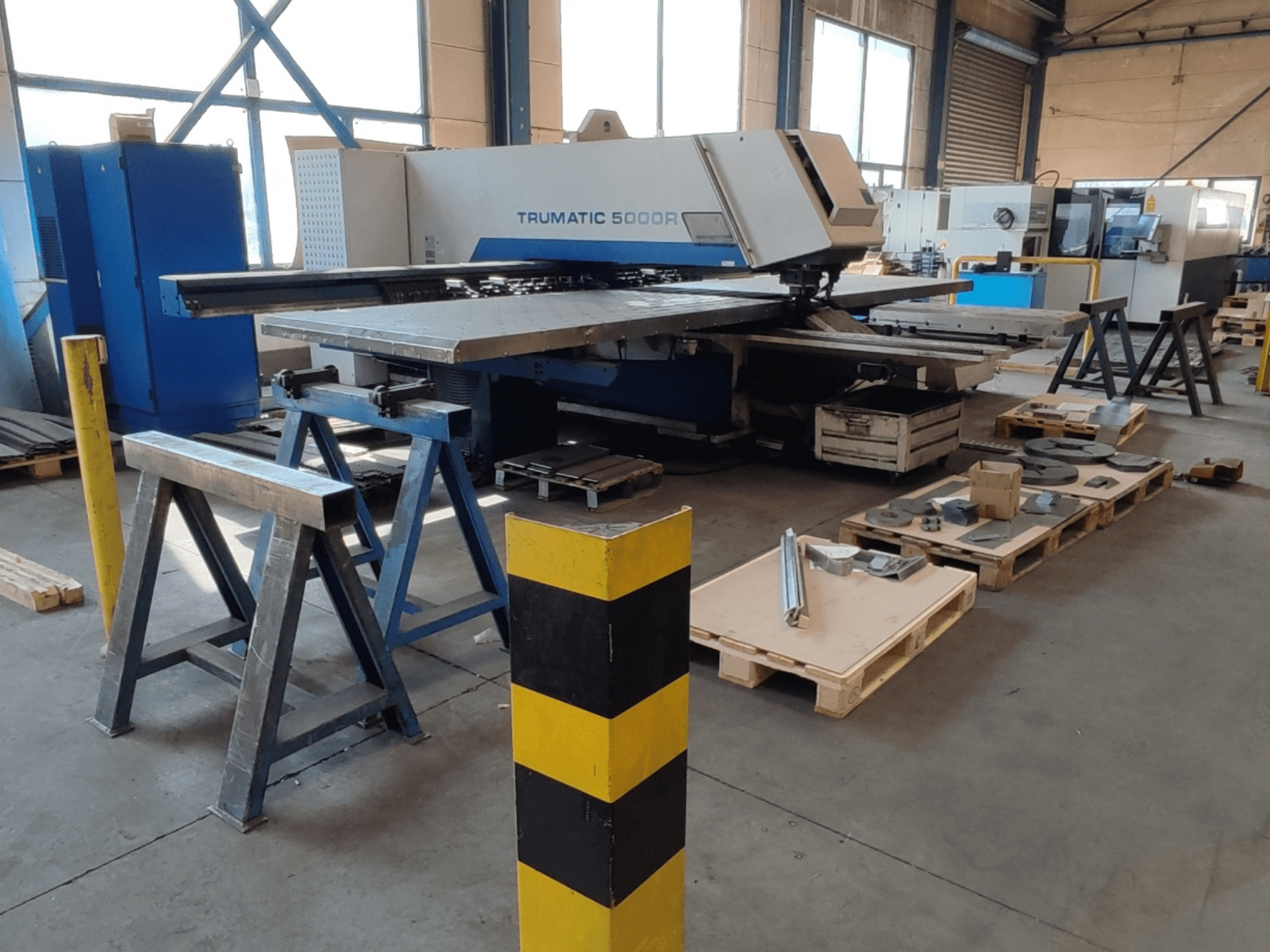 TRUMPF Trumatic 5000 R CNC perforēšanas mašīna darbnīcā, skats no sāniem ar instrumentiem un materiāliem uz paliktņiem blakus.