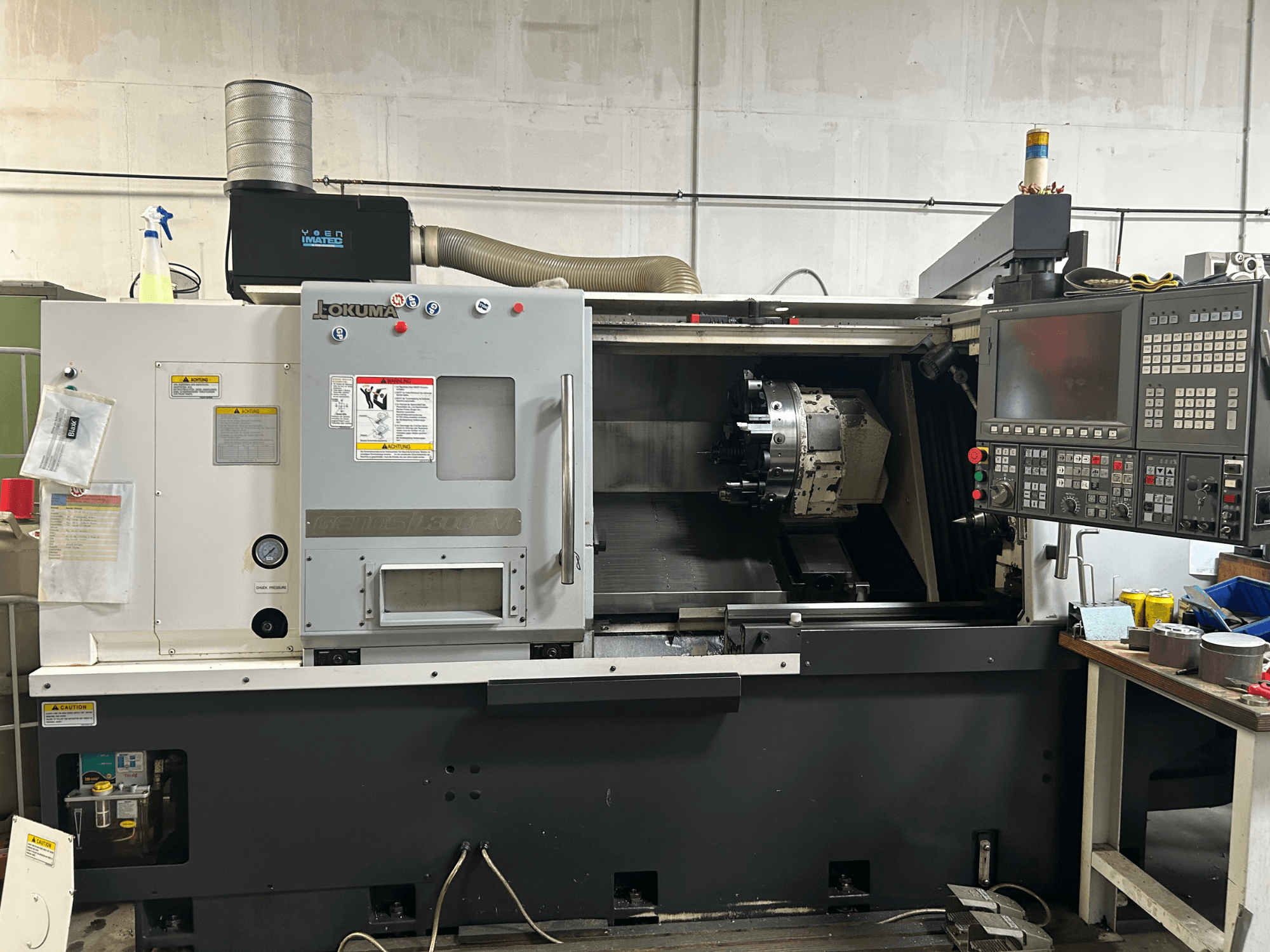 Okuma Genos L300E-M horizontālās virpošanas mašīnas priekšējais skats ar vadības paneli, redzamo vārpstu un dažādiem iestatīšanas instrumentiem.