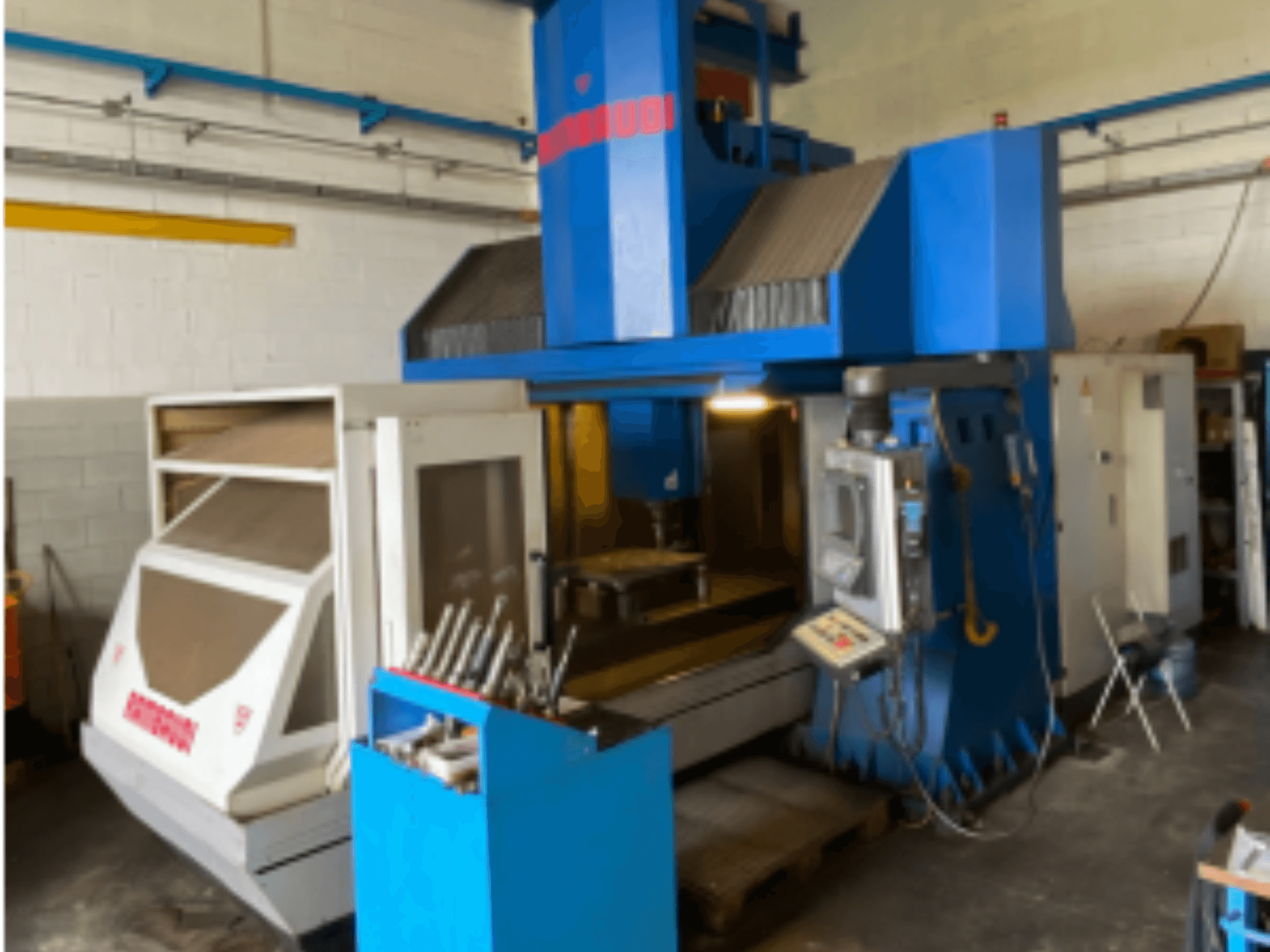 Vertikālais apstrādes centrs Rambaudi RF103, priekšējais skats, kurā redzamas vadības ierīces, instrumentu glabāšanas vieta un apstrādes zona. Zilā un baltā ārējā apdare.