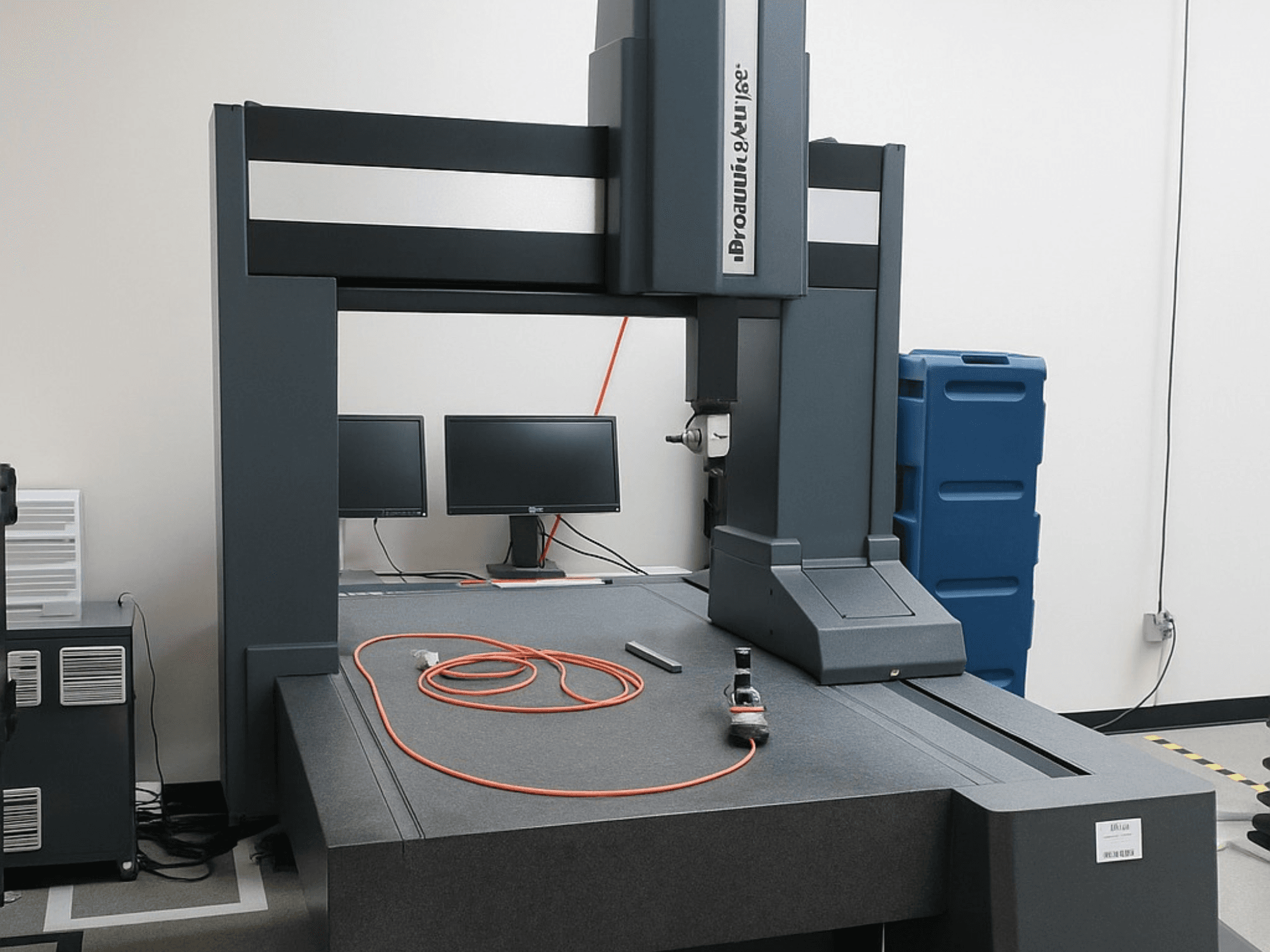 Brooks Automation koordinātu mērīšanas iekārta ar diviem monitoriem, skats no priekšpuses, ar pelēku galda virsmu un mērinstrumentiem.