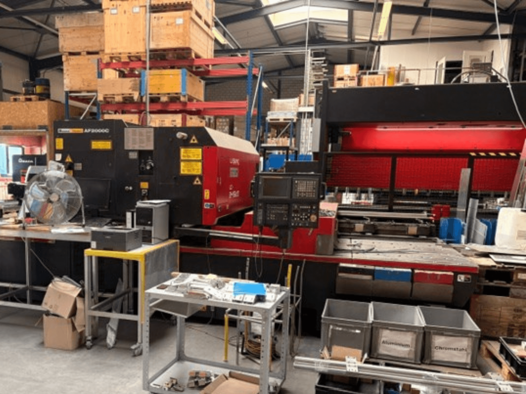 AMADA LC-2415αIII CO₂ lāzergriešanas iekārta darbnīcas vidē, kur redzams priekšskats, vadības panelis un darba zona.