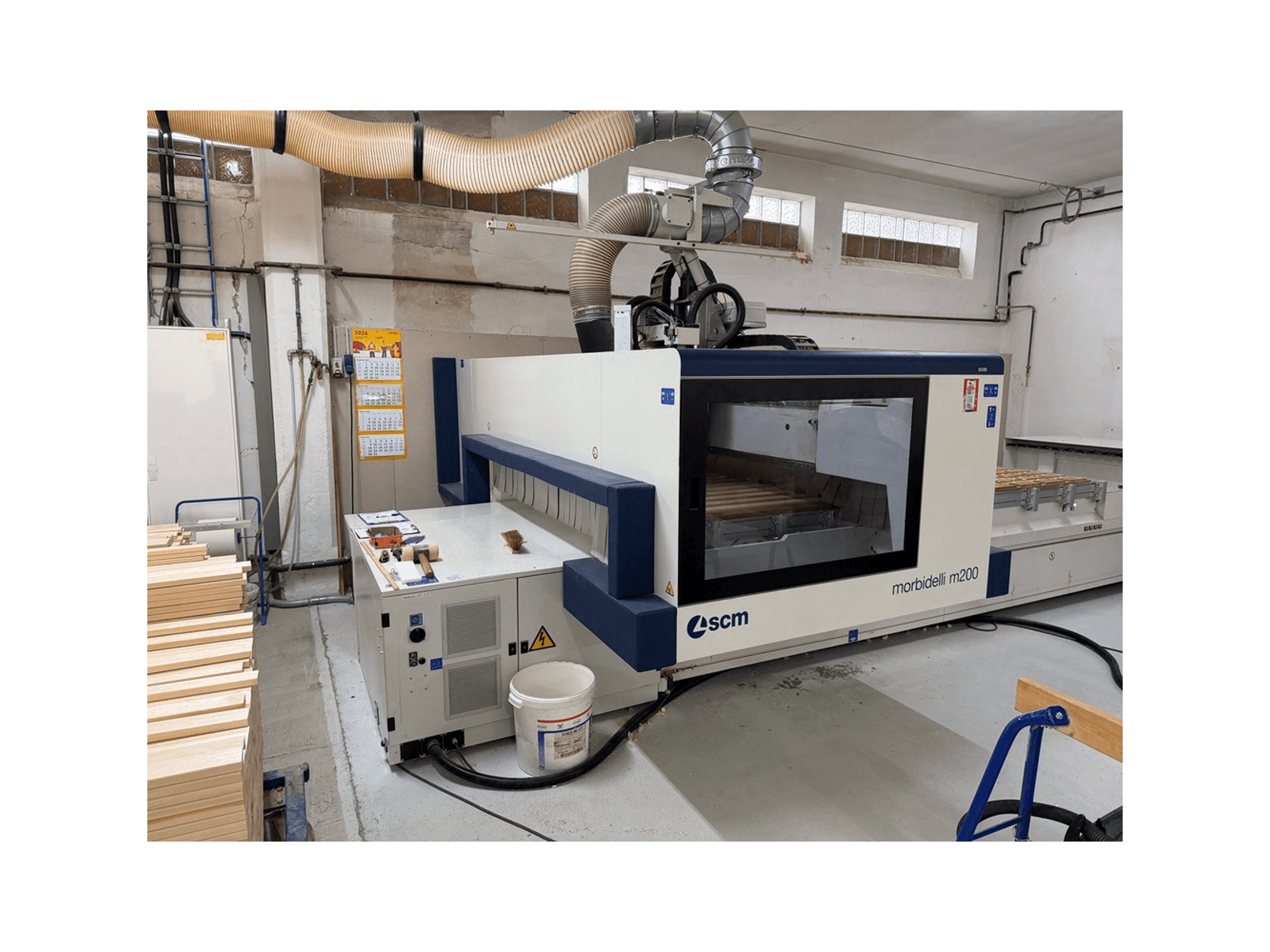 SCM Morbidelli M200F CNC apstrādes centrs darbnīcā, skats no sāniem ar koka gabaliem uz galda un ventilācijas sistēmu virs tā.