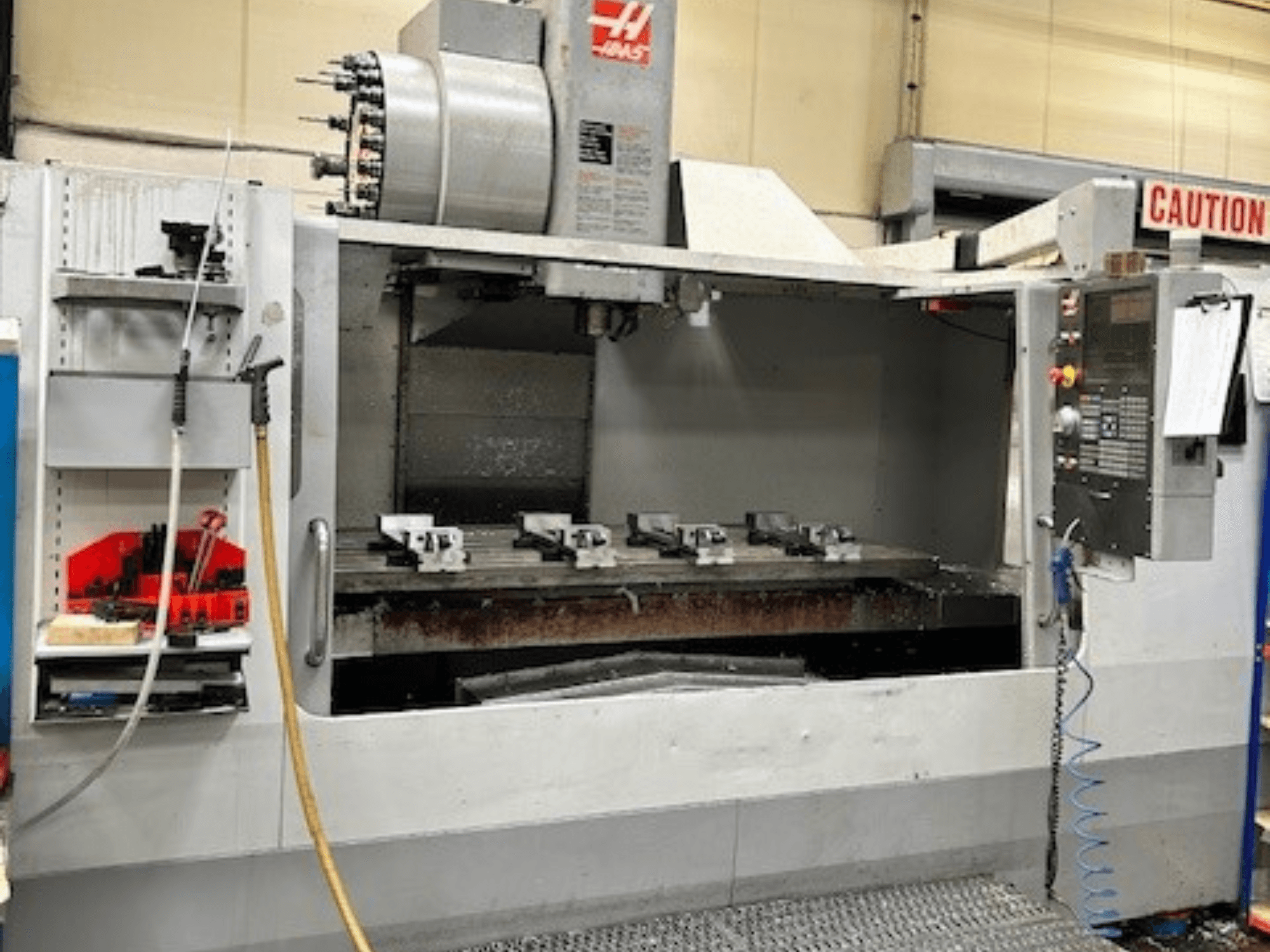 HAAS VF-9/40HE vertikālais apstrādes centrs, skats no priekšpuses, ar instrumentu turētāju un sānos redzamiem piederumiem.