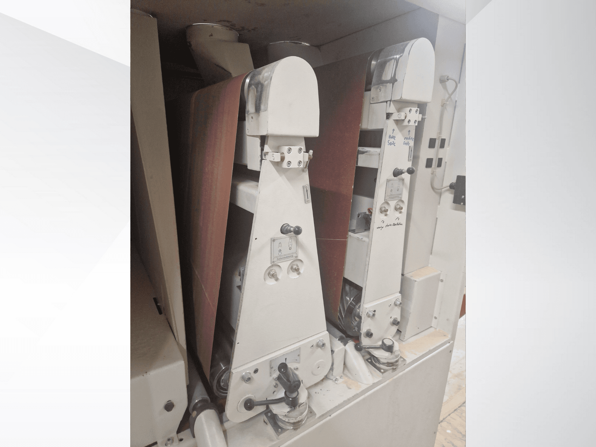 Divas rūpnieciskās slīpmašīnas ar sarkanām slīpēšanas lentēm, skats no priekšpuses, redzamas vadības ierīces un metāla detaļas.