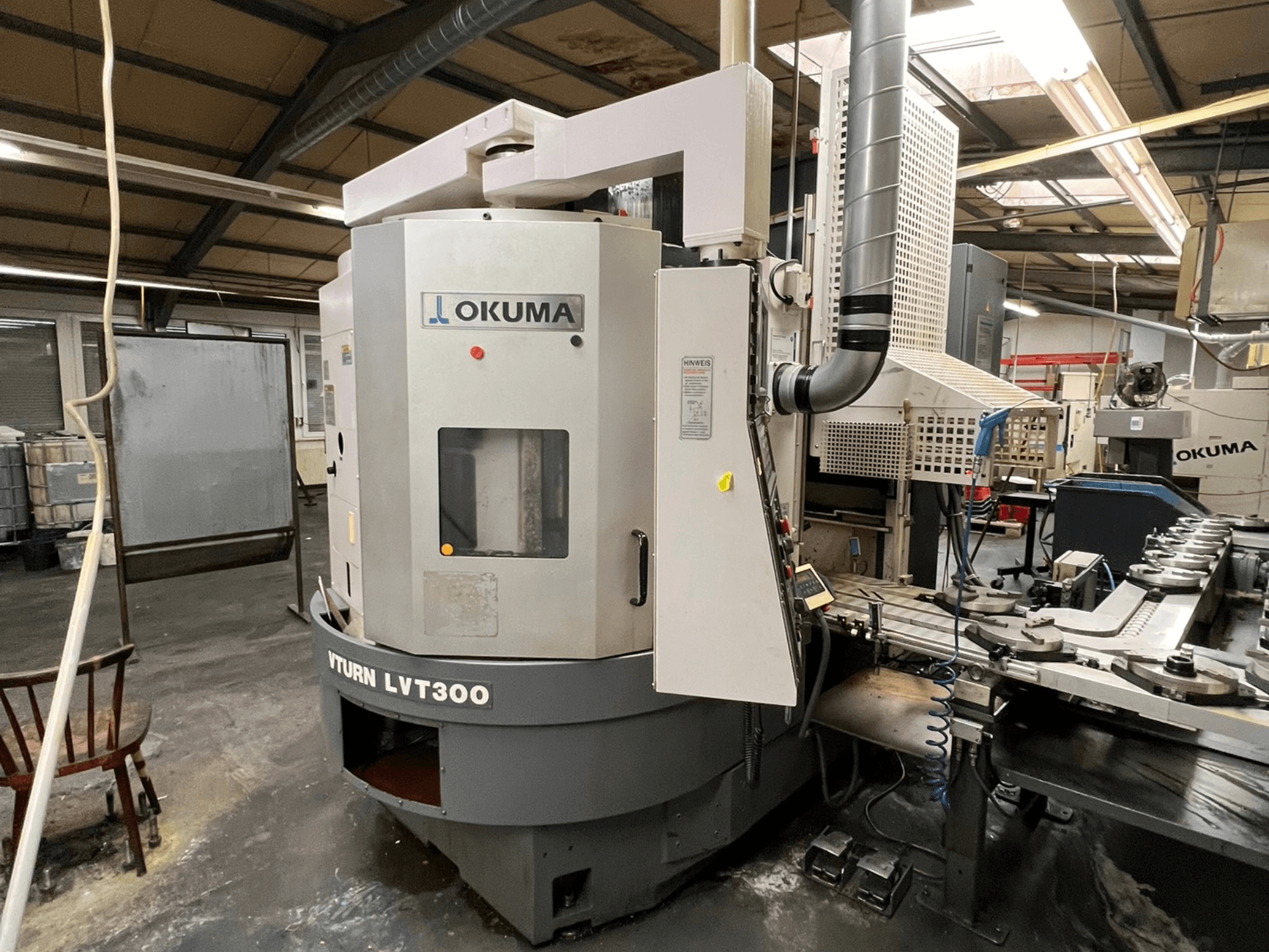 Okuma Vturn LVT300 CNC virpa, skats no sāniem rūpnieciskā vidē, ar vadības paneli un dzesēšanas šķidruma sistēmu.