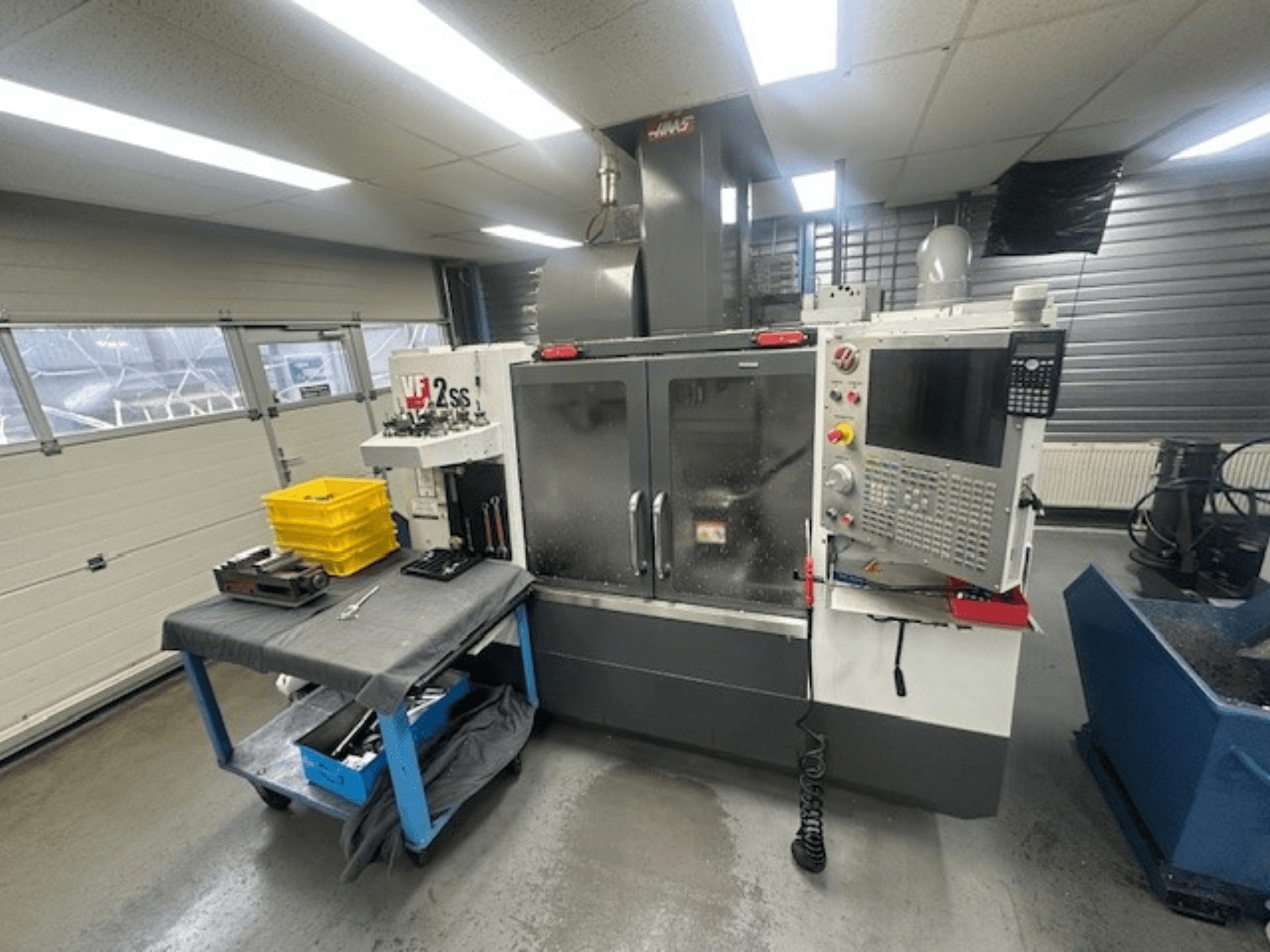 HAAS VF-2SS vertikālā apstrādes centra priekšējais skats ar vadības paneli, darba galdu un instrumentu glabāšanas staciju.