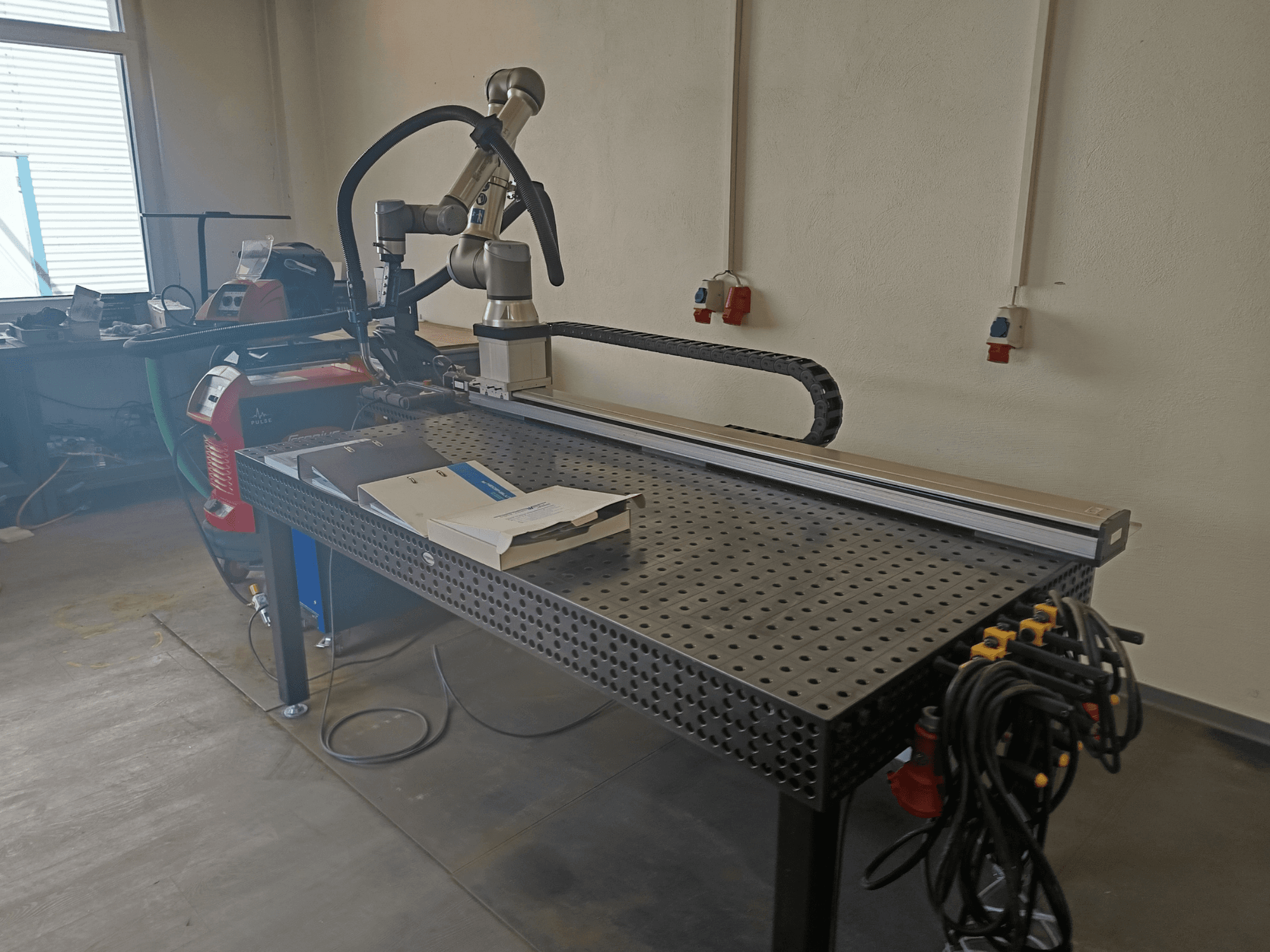 FRONIUS TransSteel 4000 impulsu metināšanas stends, skats no sāniem, kurā redzama robota roka, darba zona un strāvas pieslēgumi.