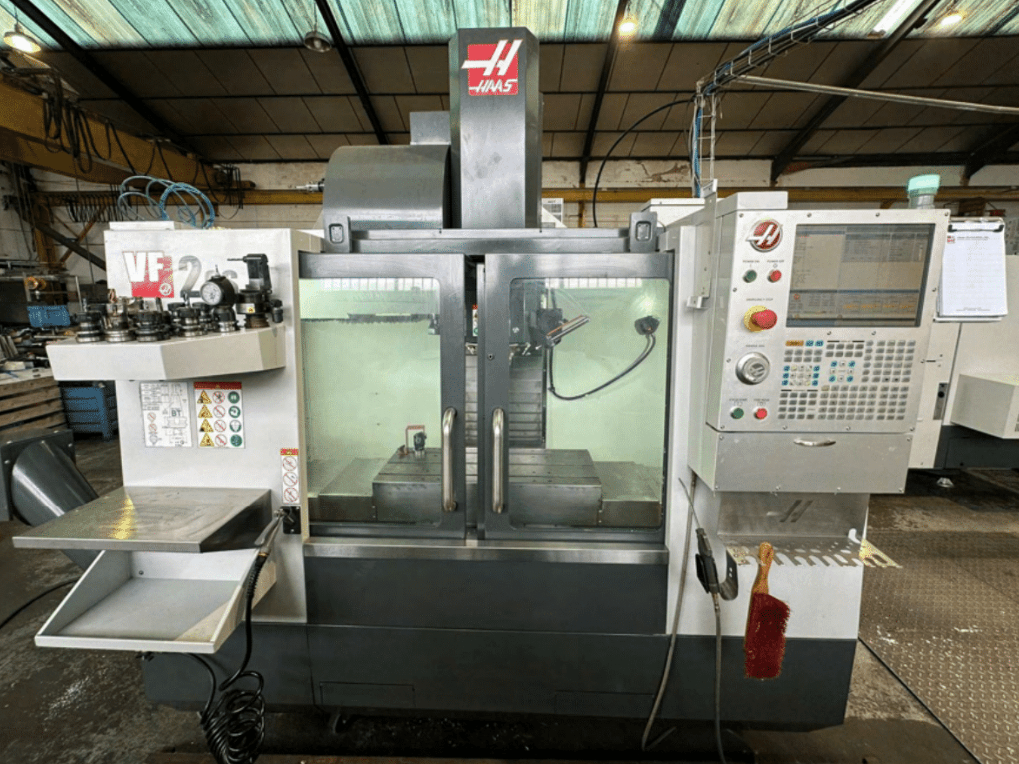 Haas VF2 CNC mašīnas priekšējais skats, kurā redzams vadības panelis, instrumentu turētāji un darba vieta ar drošības uzlīmēm un suku.