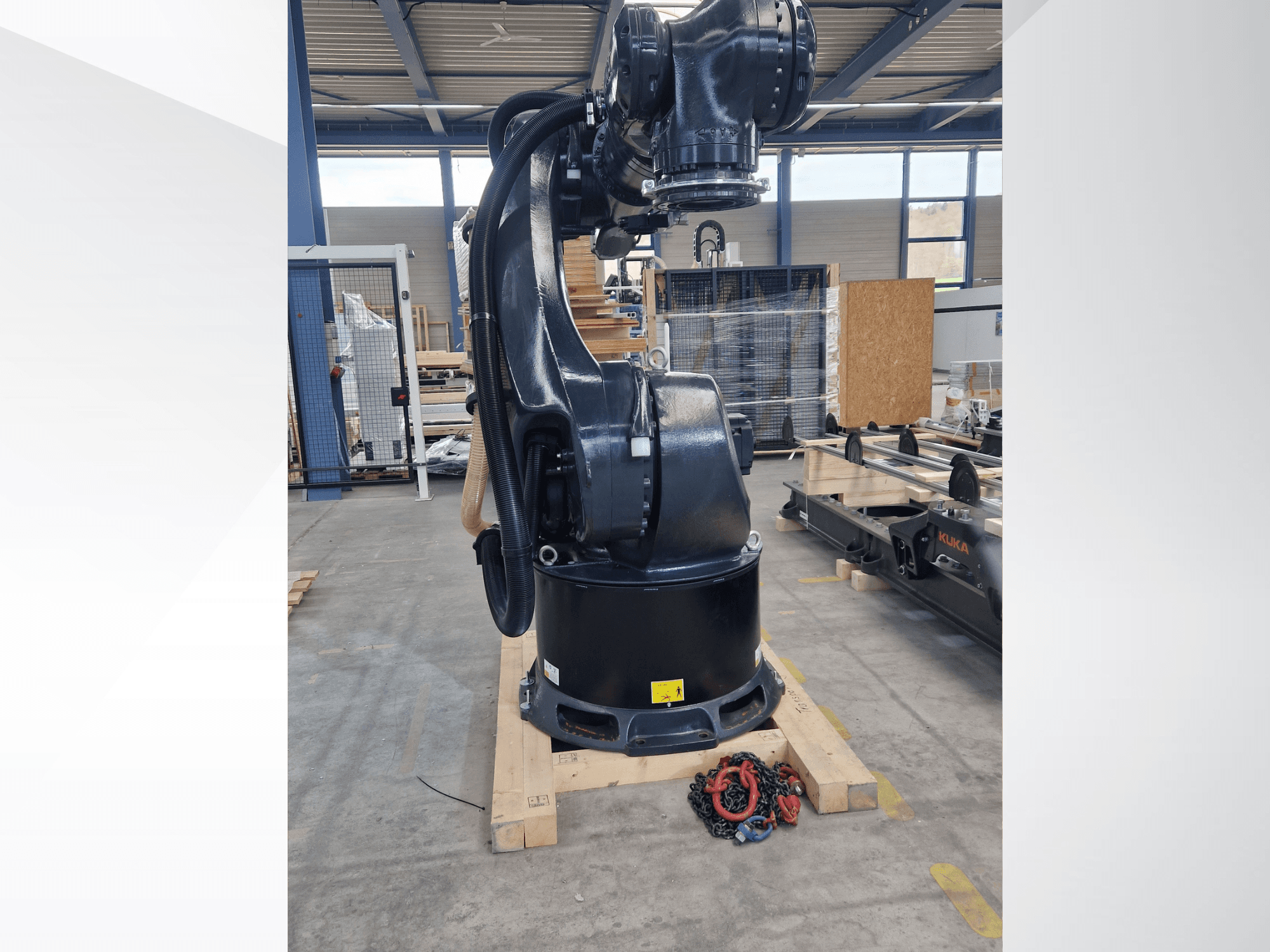 KUKA KR 340 R3330 robota roka noliktavā, sānskats, kurā redzama tās pamatne un vadi, priekšplānā — ķēdes.