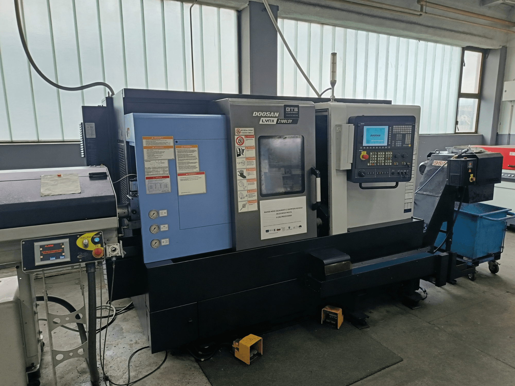 DOOSAN LYNX 2100 LSY virpošanas frēzēšanas centrs, sānu skats, kurā redzams vadības panelis un vadības pogas, ar redzamām informatīvām uzlīmēm.