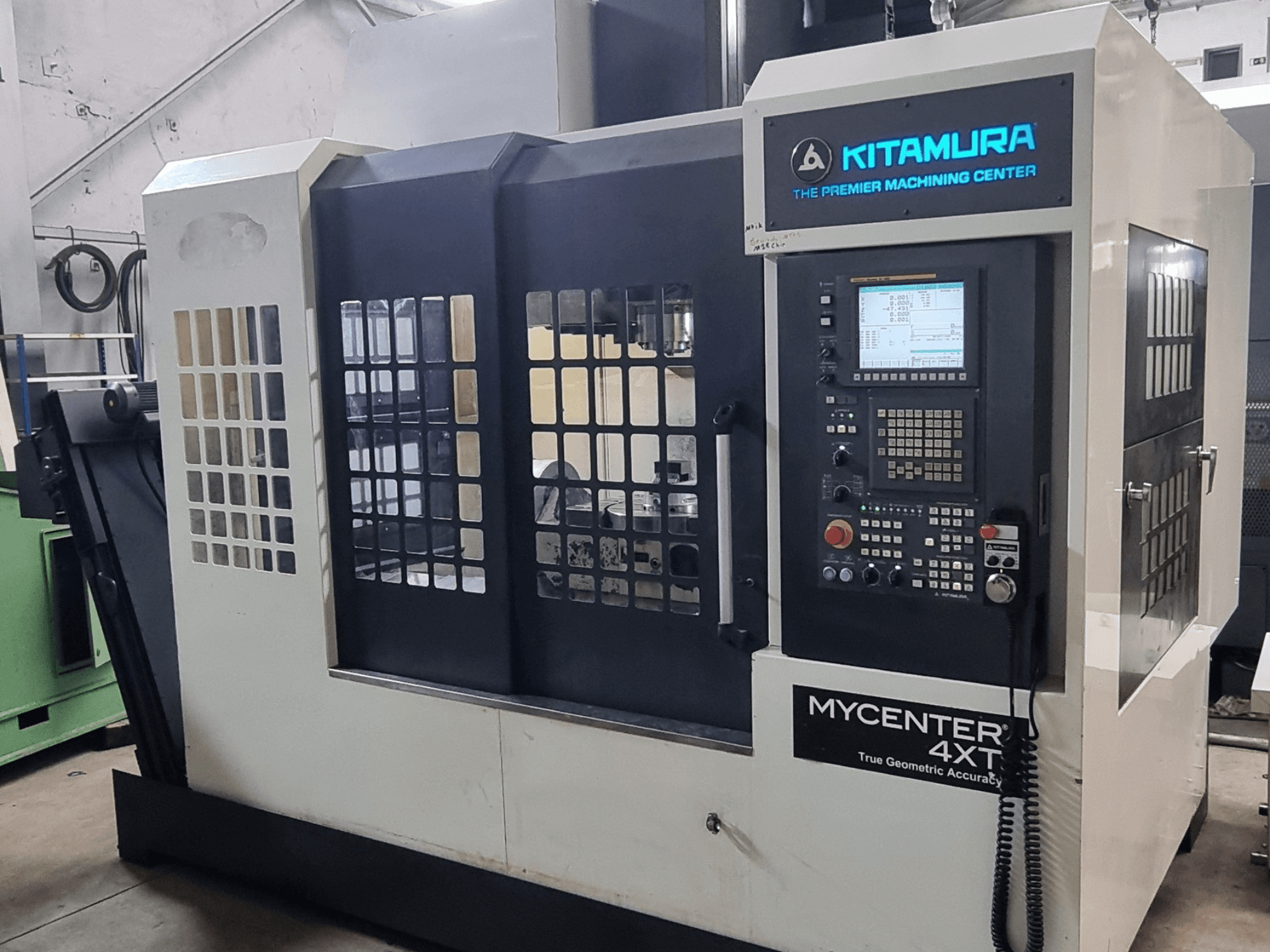 Alternatīvais teksts: Kitamura MYCENTER 4XT vertikālais apstrādes centrs, skats no priekšpuses, kurā redzams vadības panelis un mašīnas detaļas darbnīcas vidē.