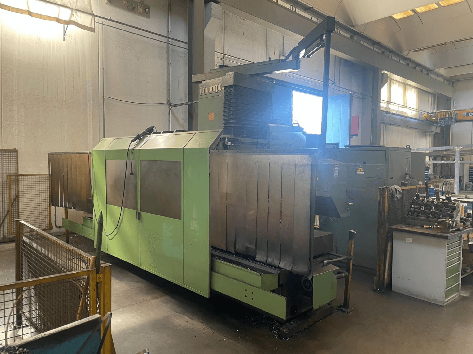 Zaļa rūpnieciska mašīna ar redzamām metāla detaļām, skats no priekšpuses; vadības panelis, drošības brīdinājumi un darba vieta blakus.