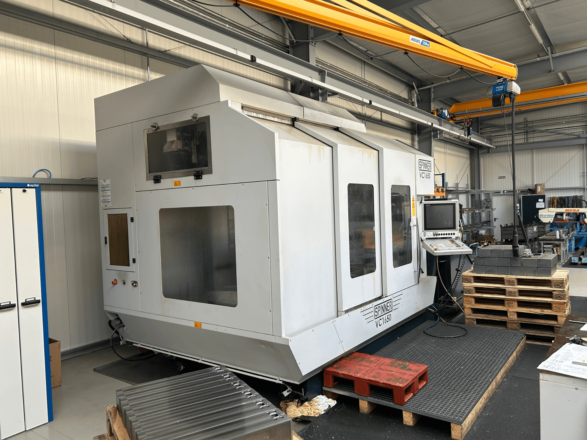 Virpļmašīna VC 1650 CNC, skats no priekšpuses, ar vadības paneli, lieliem logiem un blakus sakrautām paletēm darbnīcā.