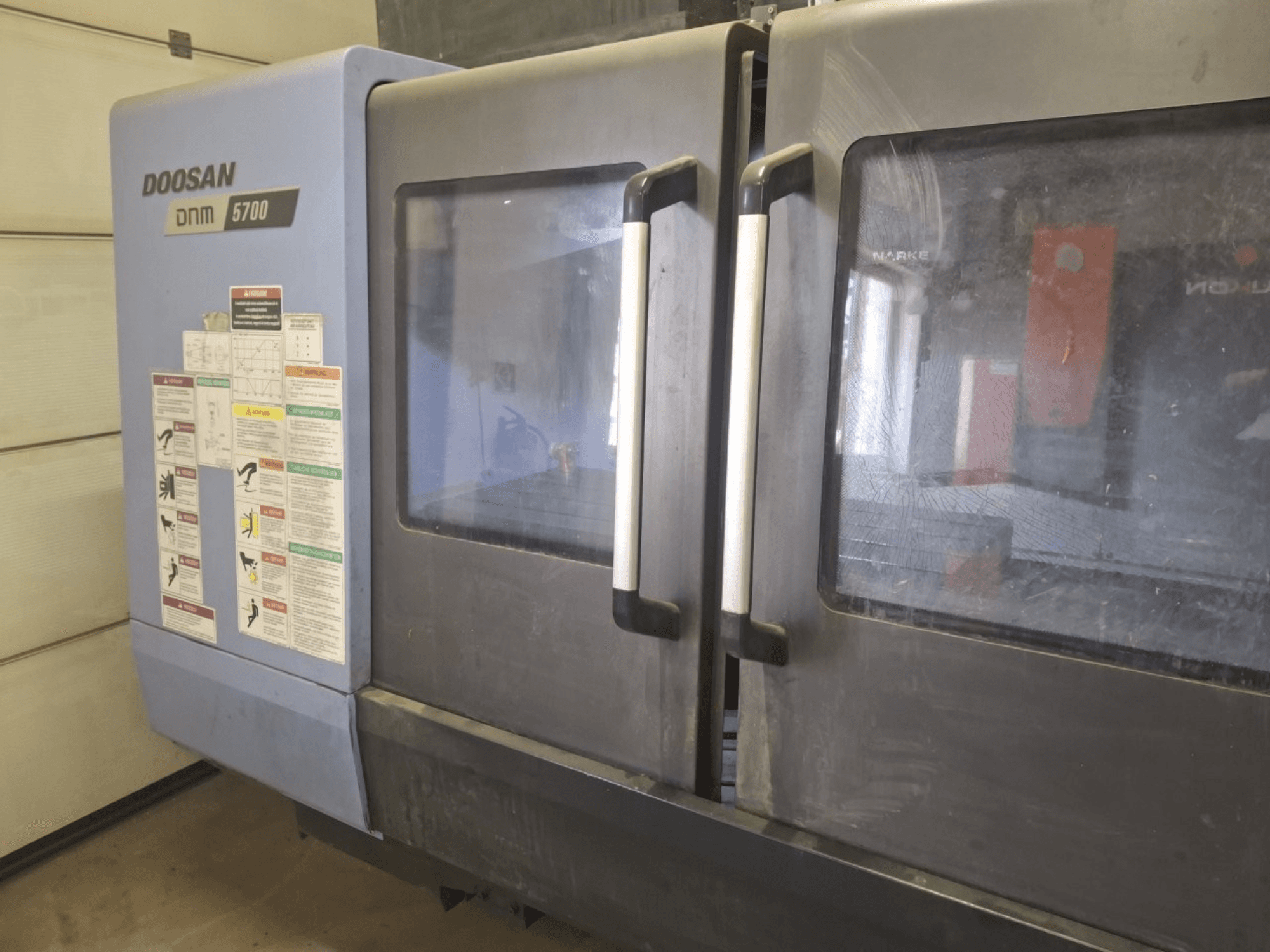 DOOSAN DNM 5700 vertikālā apstrādes centra priekšējais skats, kurā redzamas dubultās durvis un vadības paneļi ar darbības vadlīnijām.