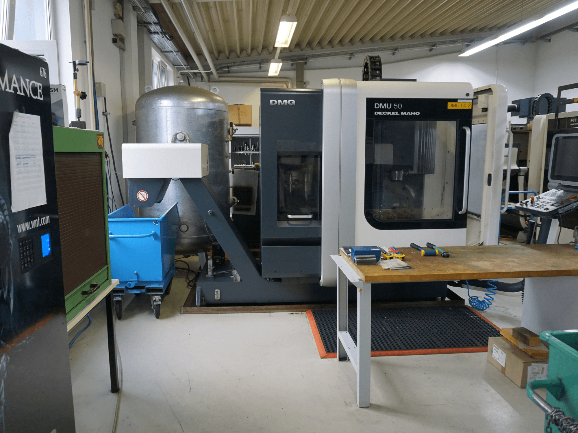 DMG DMU 50 frēzmašīna darbnīcā, skats no priekšpuses, ar instrumentiem uz koka galda un zilu atkritumu tvertni uz riteņiem blakus.