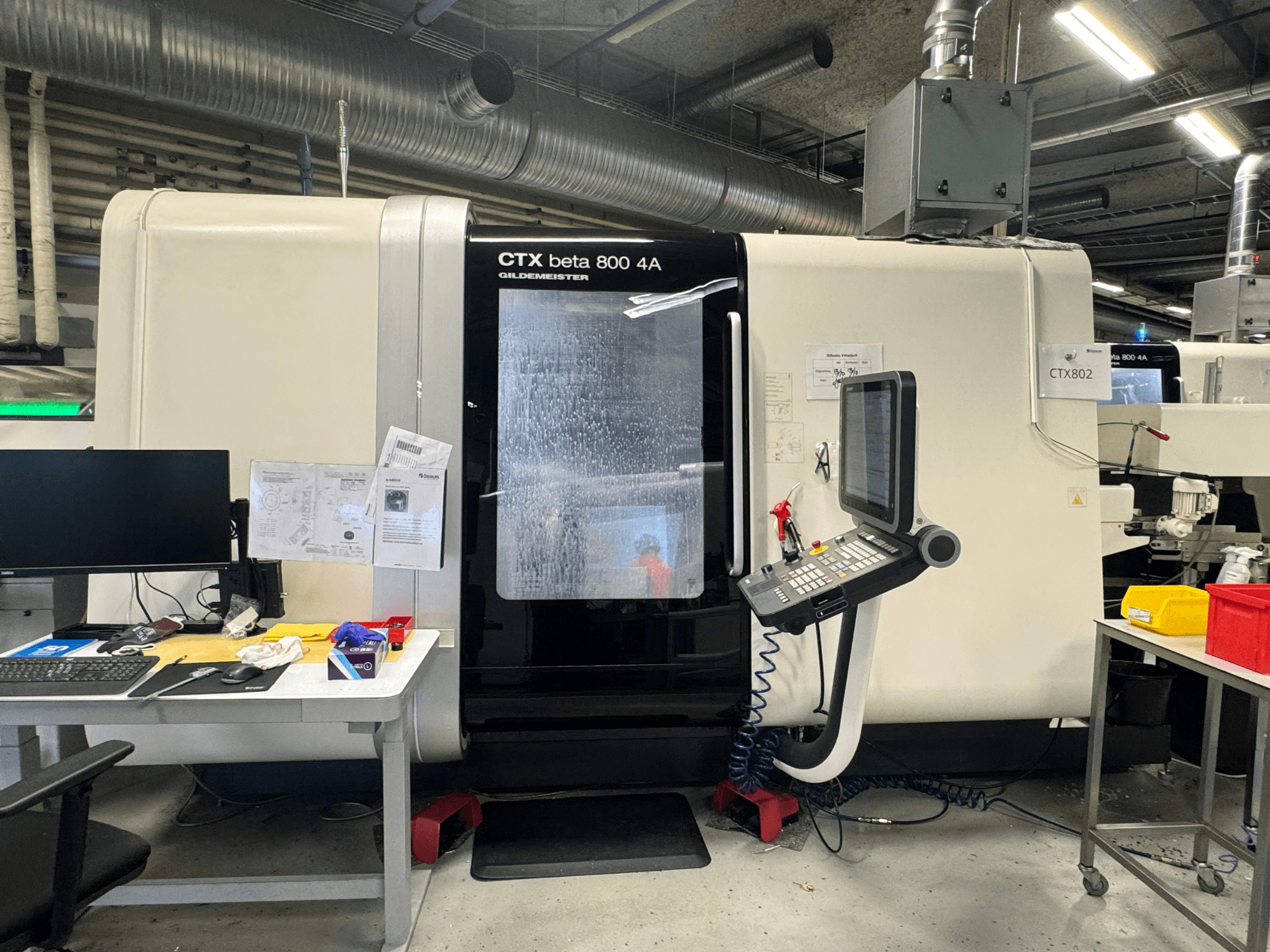 CTX beta 800 4A CNC mašīna darbnīcā, skats no sāniem, kurā redzams vadības panelis un darba vieta, kas pārblīvēta ar instrumentiem un dokumentiem.