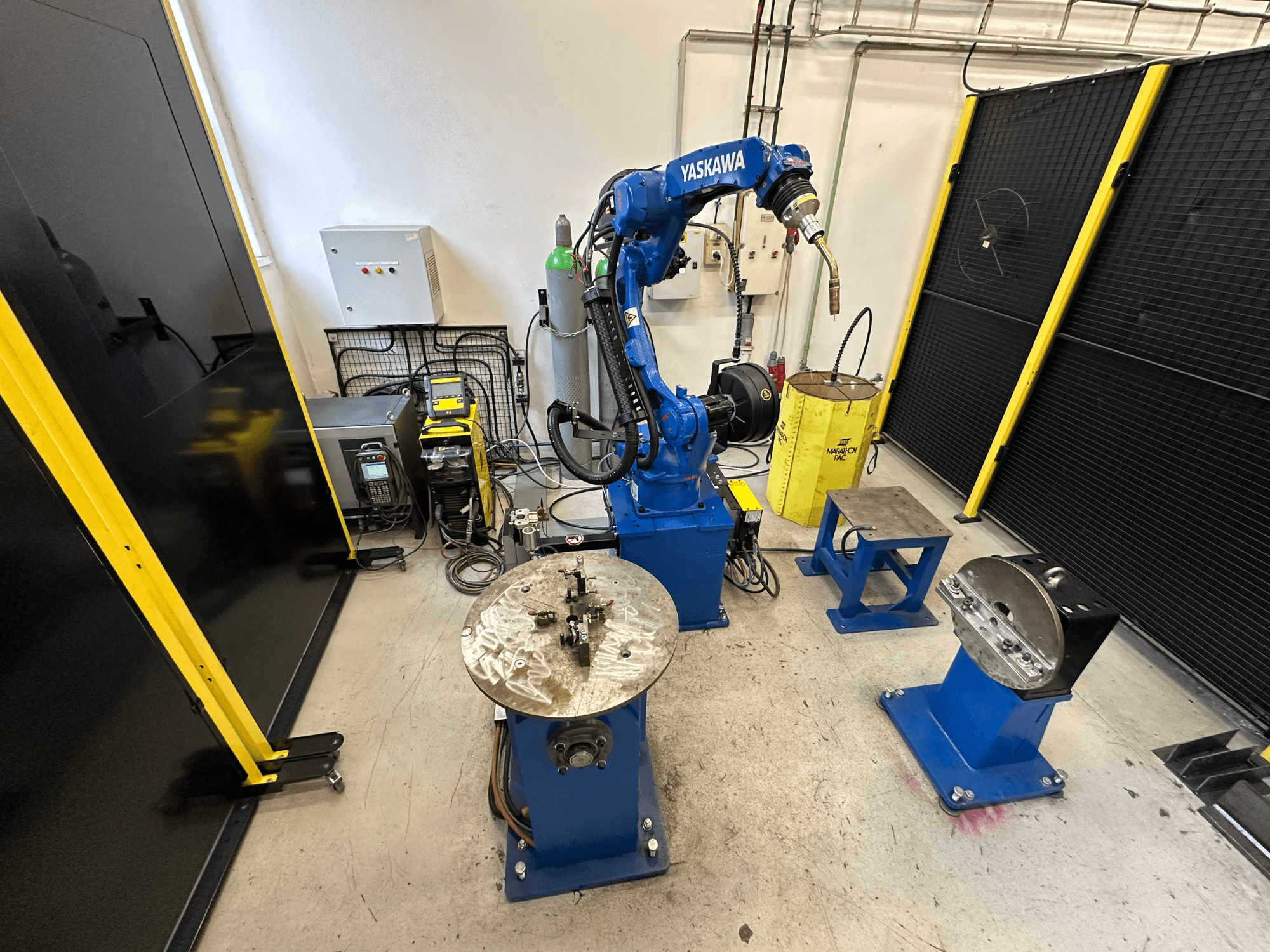 Yaskawa robotizētā roka zilā krāsā ar diviem darba galdiem, metināšanas aprīkojumu un drošības barjerām; rūpnieciskās darba vietas skats no priekšpuses.