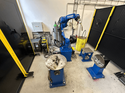 Yaskawa robotizētā roka zilā krāsā ar diviem darba galdiem, metināšanas aprīkojumu un drošības barjerām; rūpnieciskās darba vietas skats no priekšpuses.