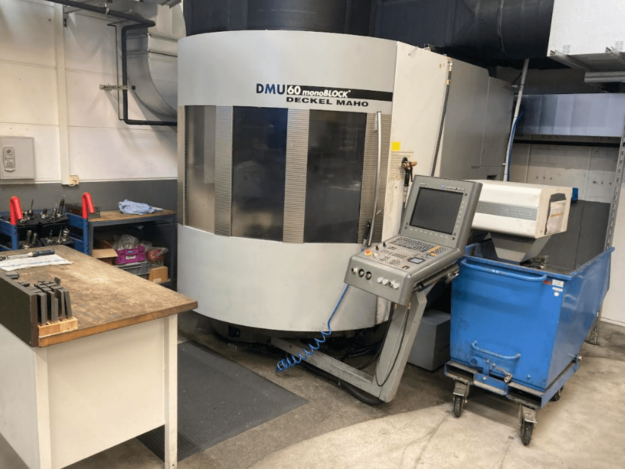 DMG MORI DMU 60 Monobloka vertikālais apstrādes centrs, priekšējais skats ar vadības paneli, darba vietu un redzamu zilu instrumentu ratiņu.