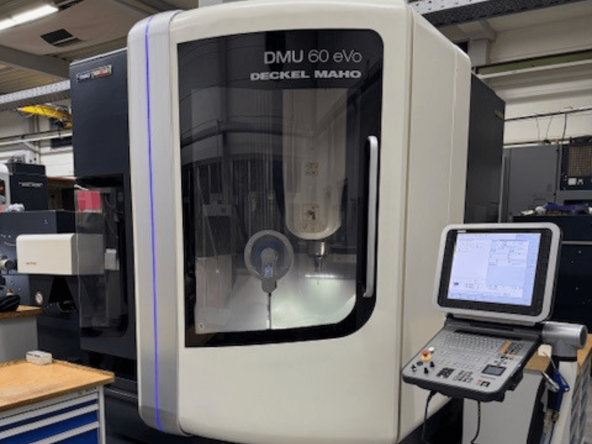 DMU 60 eVo, DECKEL MAHO, priekšējais skats uz CNC frēzmašīnu ar vadības paneli un displeja ekrānu, uz kura redzamas precīzas detaļas.