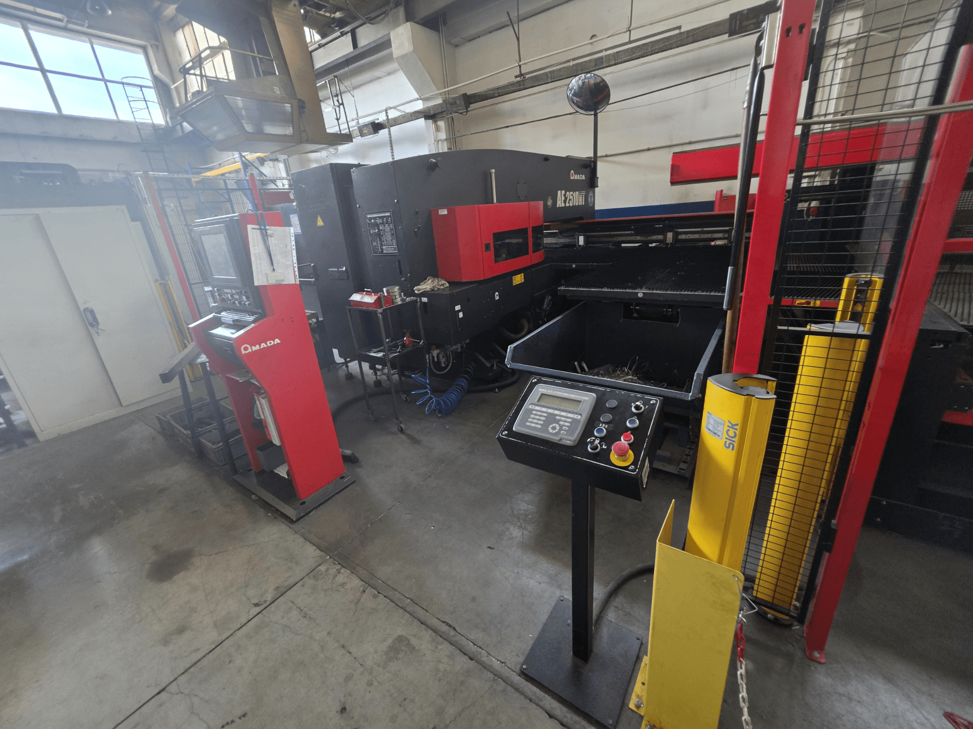 CNC perforācijas mašīna AMADA AE-2510NT, skats no priekšpuses ar vadības paneli, drošības elementiem un apkārtējo rūpniecisko vidi.