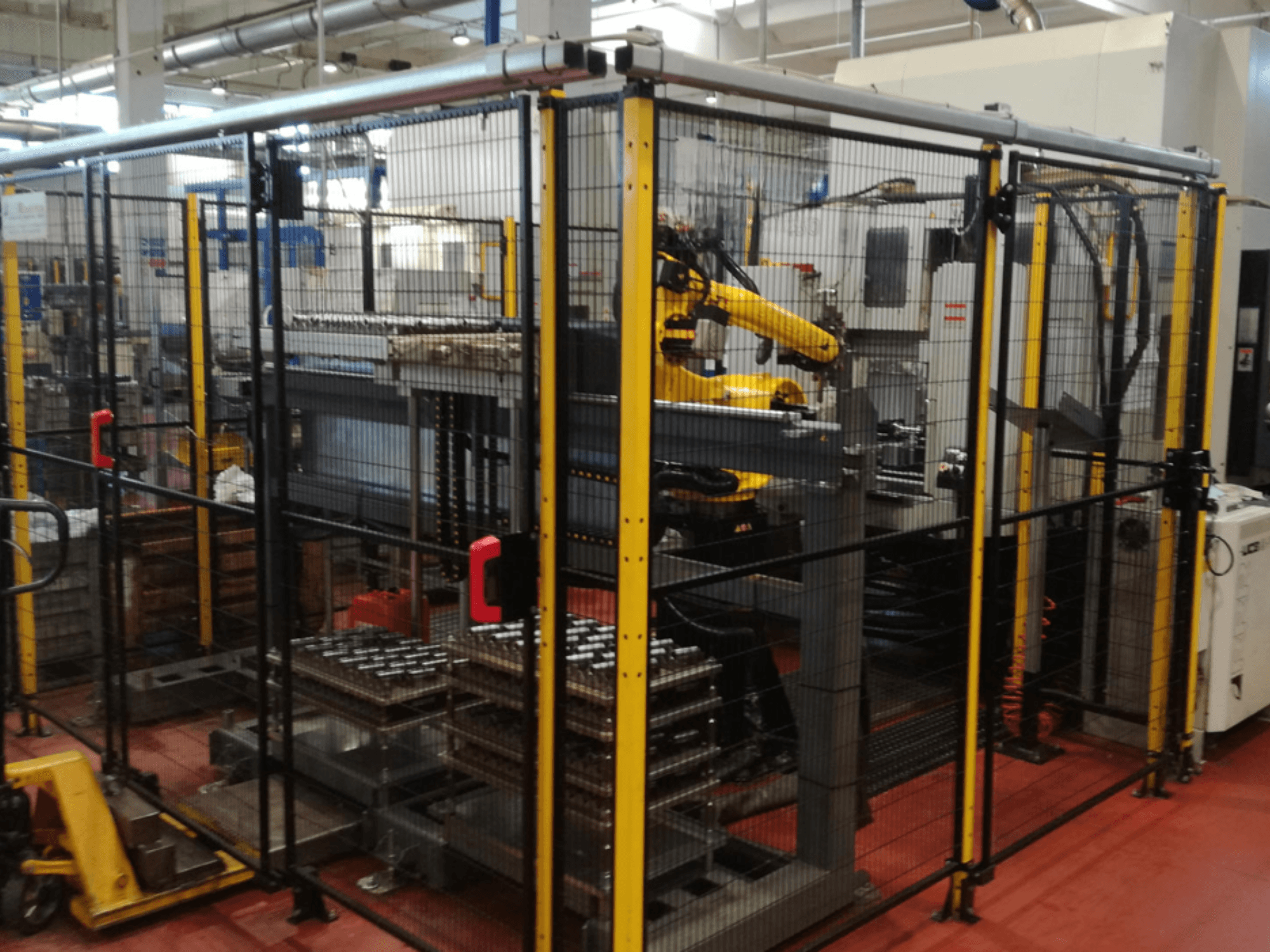 Vertikālais apstrādes centrs Enshu EV450 ar robotizētām rokām un vairākām kārtām novietotām detaļām, iežogotā darba telpā. Skats no priekšpuses.